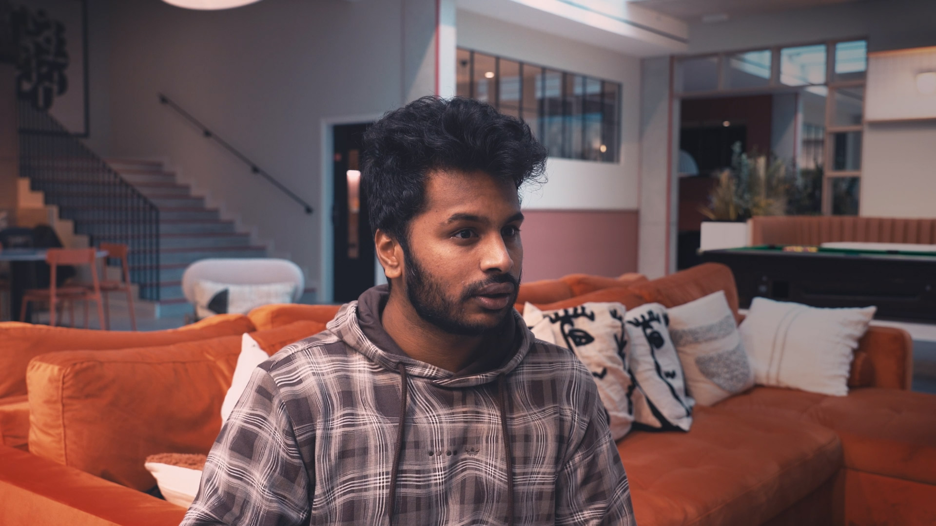 A person with dark hair and a beard sits on an orange sofa in student accommodation in Coventry, wearing a plaid hoodie. Behind them is a modern living space with a staircase, large windows, and patterned cushions. The lighting is soft and natural.