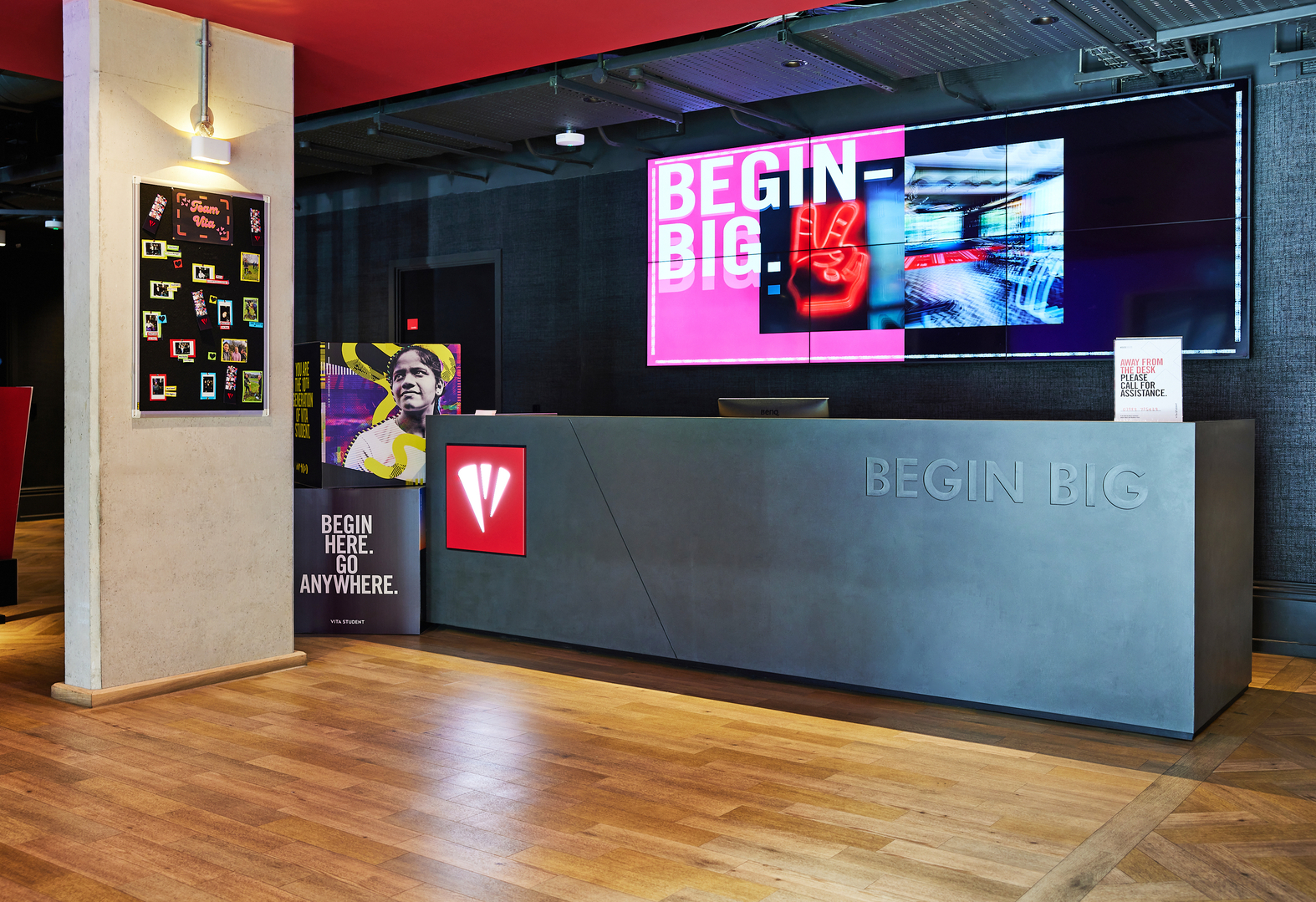 Reception area with a black desk featuring "BEGIN BIG" text. Behind the desk, large screens display abstract graphics. To the left, a poster with a person and the phrase "BEGIN HERE. GO ANYWHERE." adorns the wall on a wooden floor.