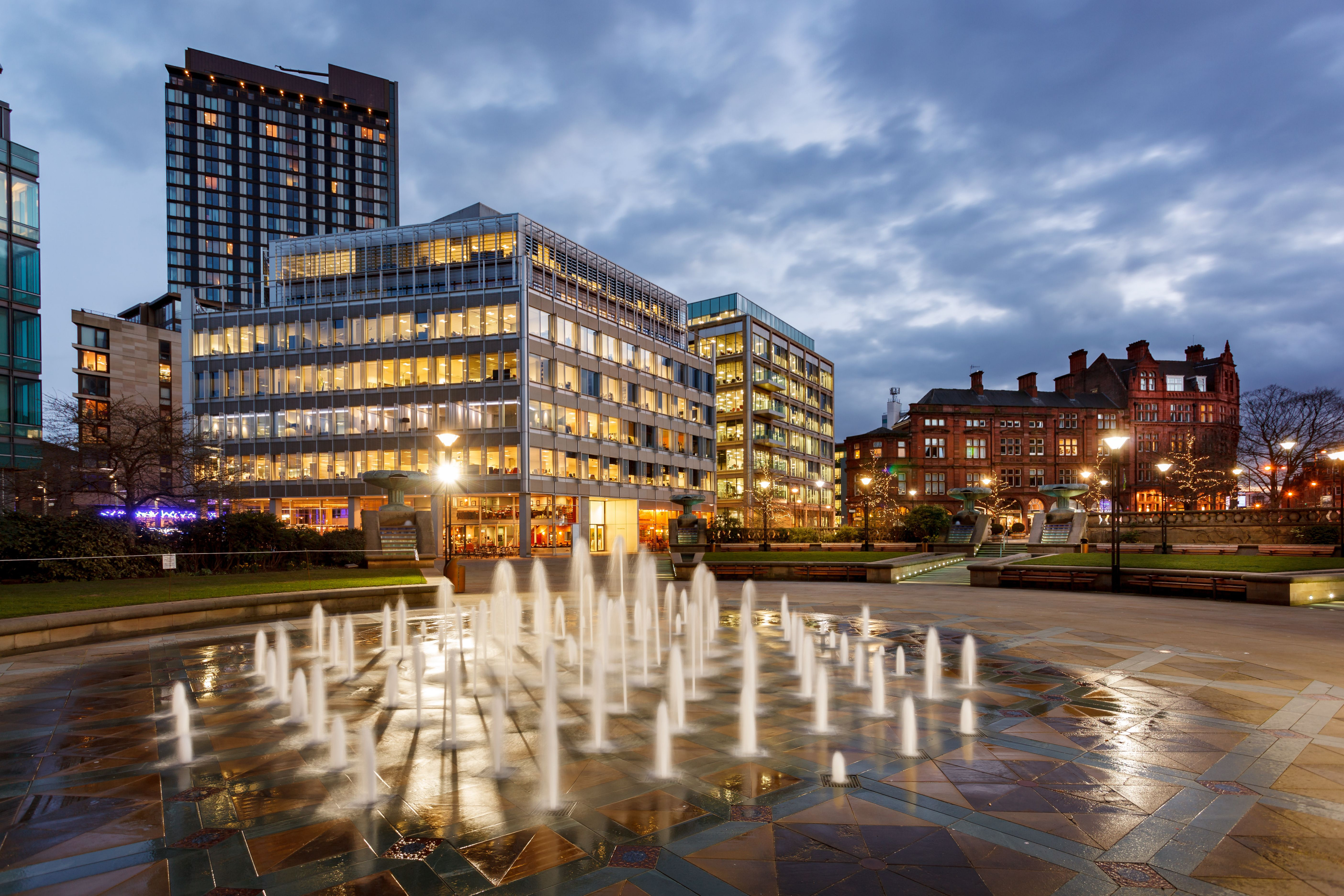 A modern city plaza at dusk, featuring an illuminated fountain in the foreground. Surrounding the plaza are contemporary glass buildings and an older red-brick structure under a cloudy, evening sky.