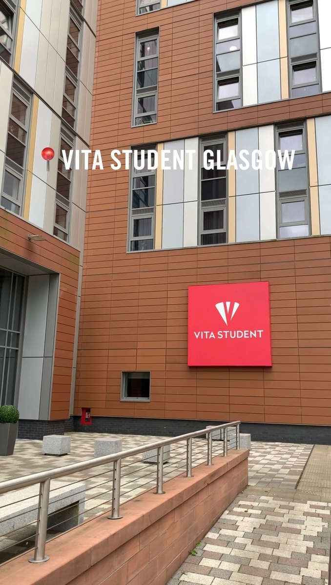 The image shows the exterior of the Vita Student accommodation in Glasgow, featuring a modern building with orange and beige panels. A large red sign displays the Vita Student logo. The courtyard has a ramp and potted plants.