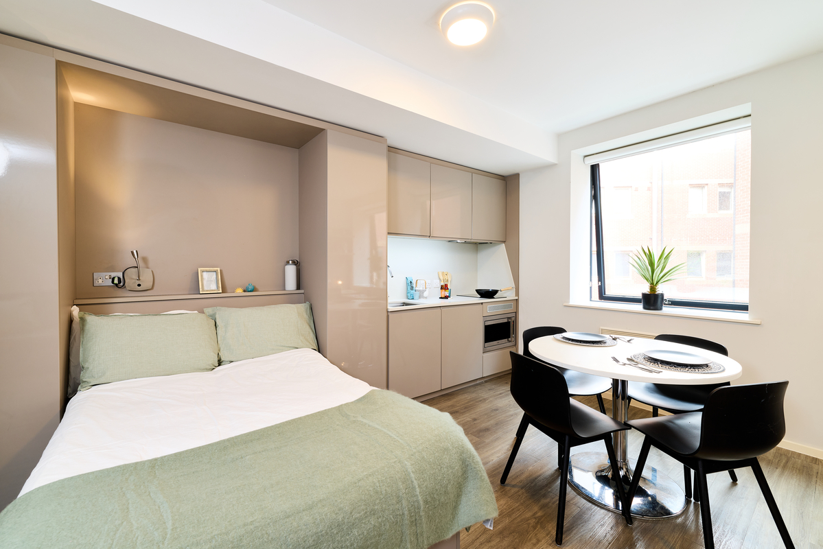Modern studio apartment in Portland House featuring a bed, small kitchen area, round dining table with four black chairs, a window with a plant on the sill, and minimalistic decor.