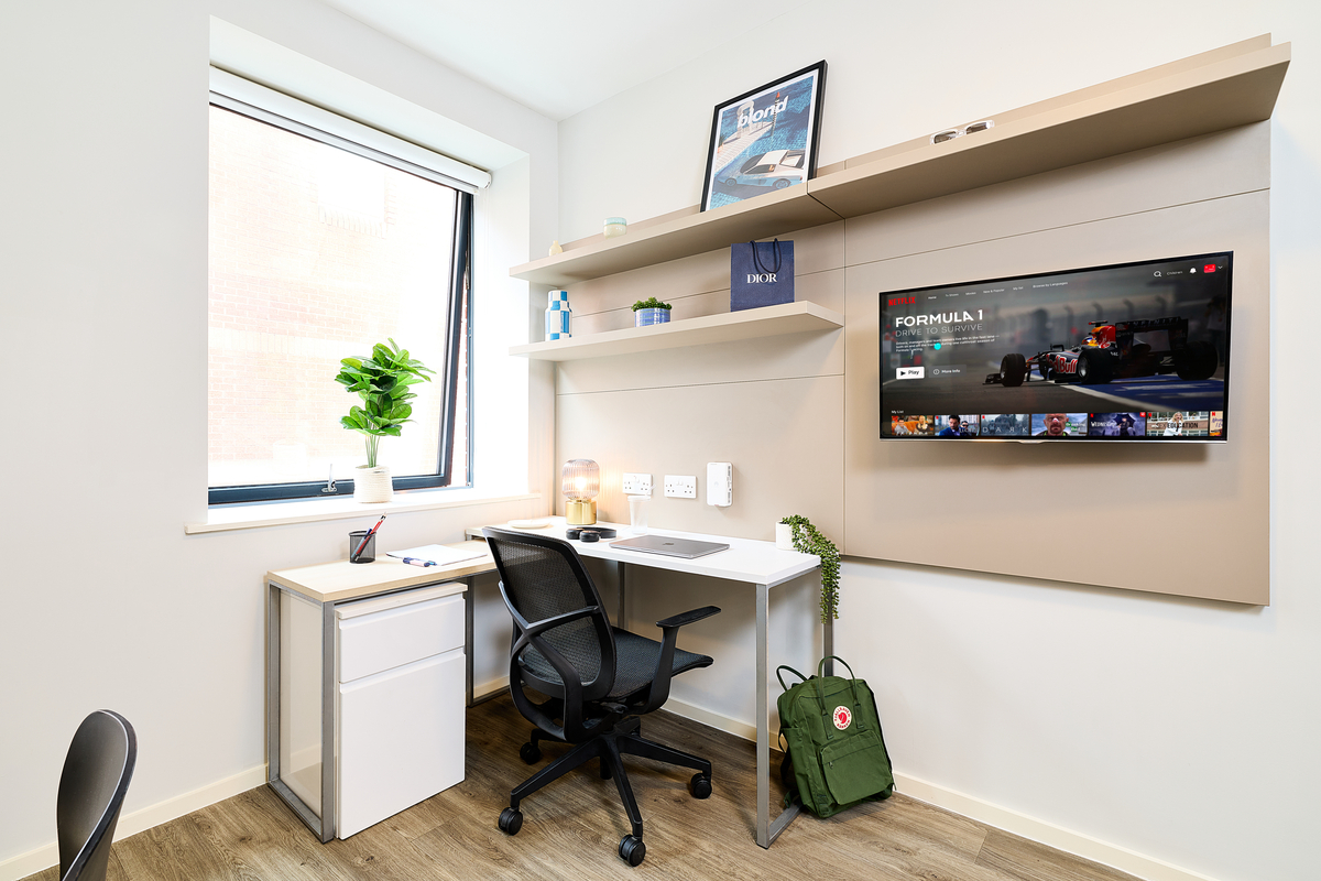 A modern, tidy study area in Portland House features a desk, office chair, shelves, TV displaying a Formula 1 race, a window with a potted plant, and a green backpack on the floor beside the desk.