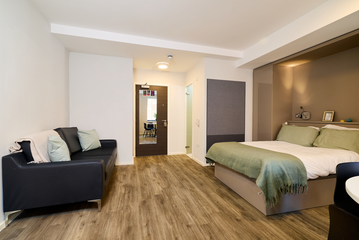Modern studio apartment at Portland House with wood flooring, a double bed with green bedding, a black sofa with cushions, and soft lighting. A doorway and desk area are visible in the background.