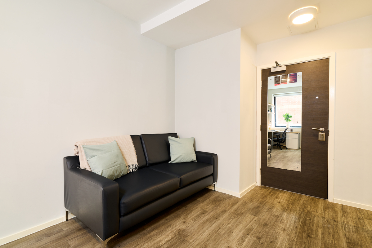 A modern, minimalist room at Portland House features a black leather sofa, pale green and beige cushions, a beige throw, wood-style flooring, and a dark wooden door with a mirror reflecting another room.