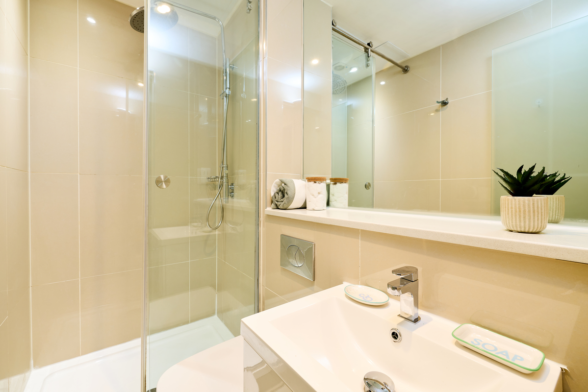 Modern bathroom at Portland House with a glass shower, beige tiled walls, white sink with soap dishes, rolled towels, jars, and a potted plant on a shelf above the toilet.