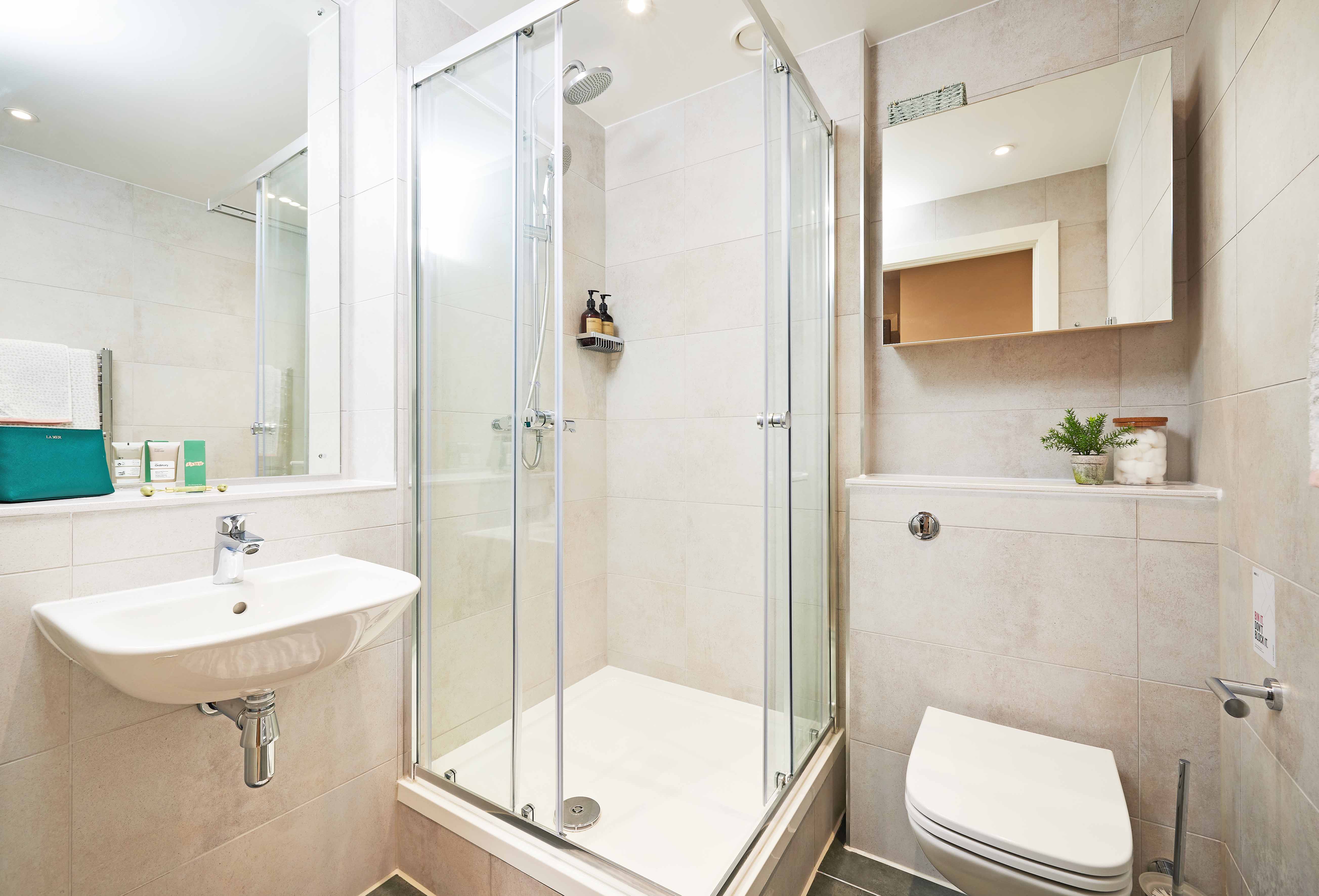 Small, modern bathroom with a glass shower enclosure, wall-mounted sink, and toilet. Light beige tiles cover the walls and floor. A mirror hangs above the sink, and a small plant decorates a shelf. Toiletries are visible in the shower.