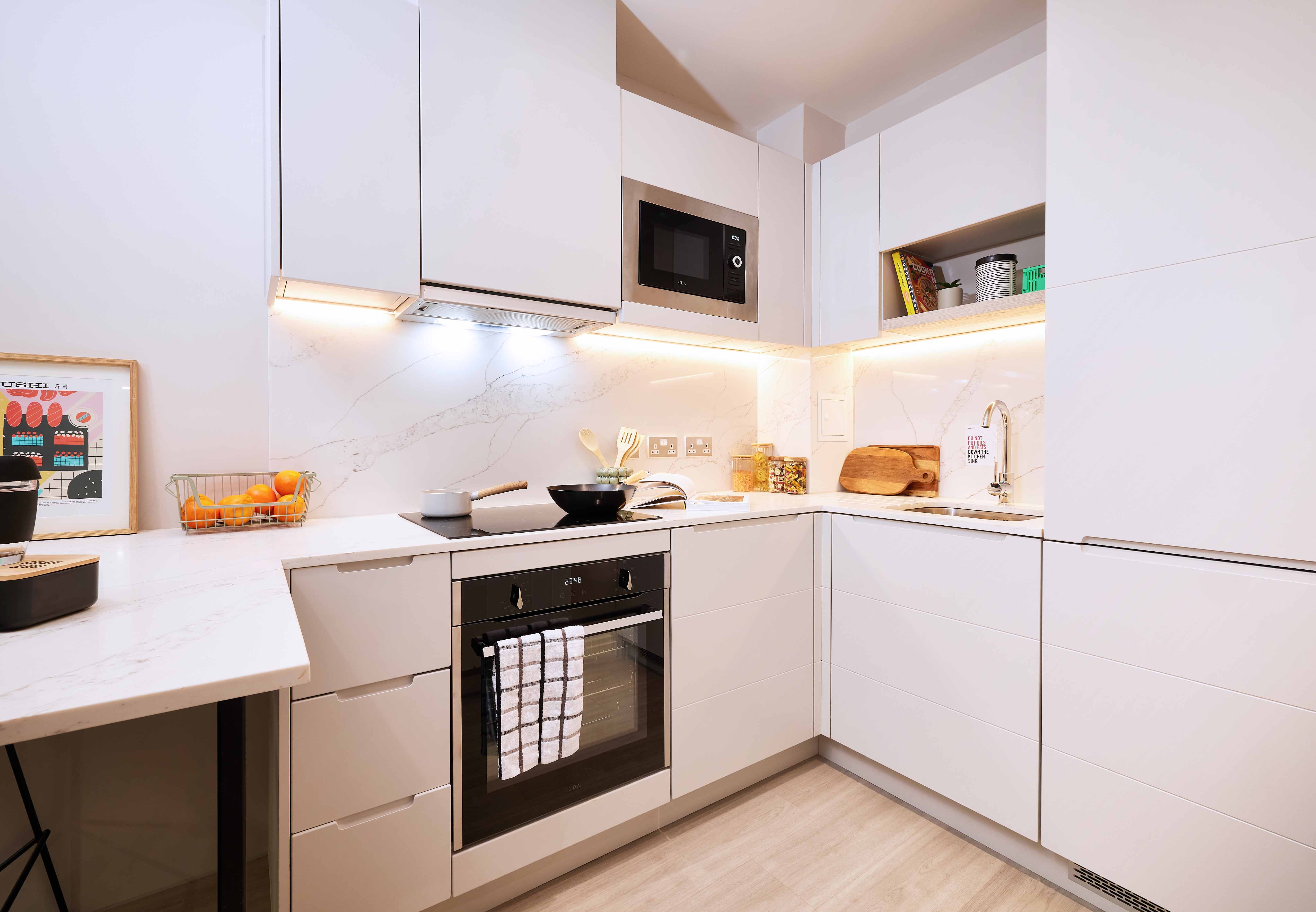 Modern kitchen with white cabinets, marble countertops, and wooden flooring. It features a built-in oven, microwave, sink, and under-cabinet lighting. A basket of fruit and framed art are on the counters.