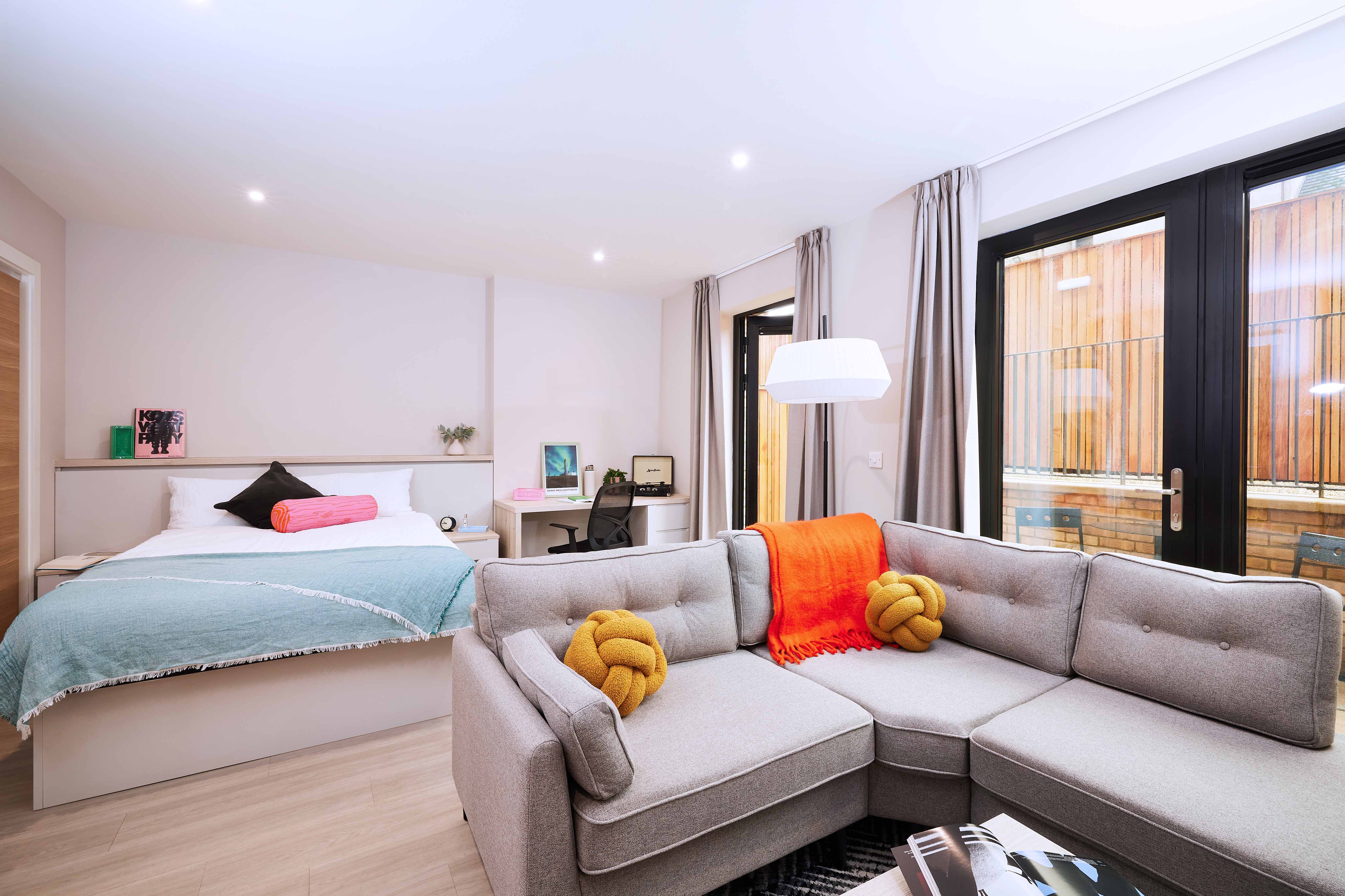 A modern studio apartment with a gray sectional sofa, two yellow knot pillows, and a red throw. A bed with blue bedding is in the background. Large windows let in natural light. A desk with a chair is in the corner. Neutral tones create a cozy vibe.