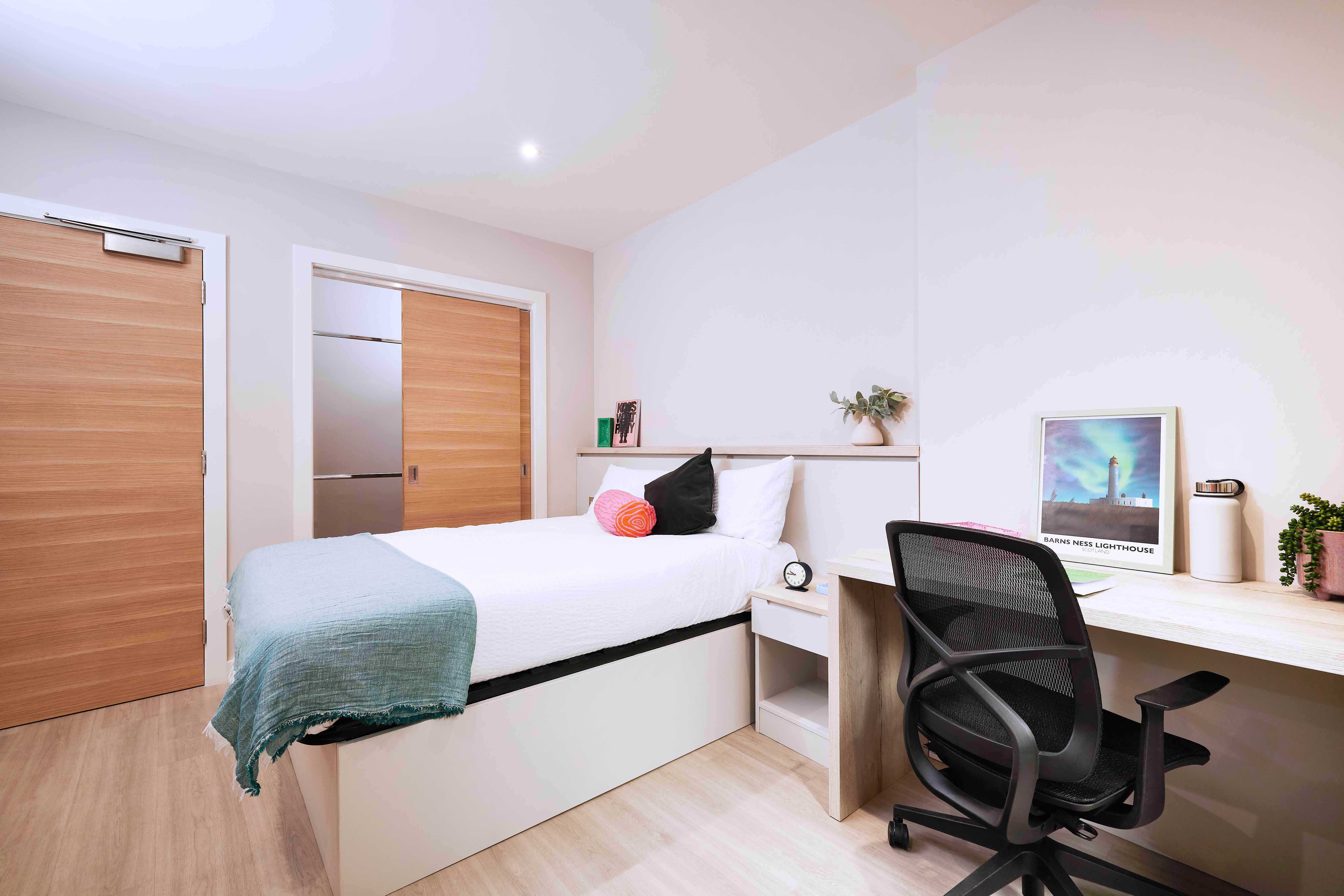 A tidy bedroom with a single bed on the left, featuring white and teal bedding and an orange cushion. On the right, there's a wooden desk with a black chair, decorated with a plant, books, a framed picture, and some stationery.