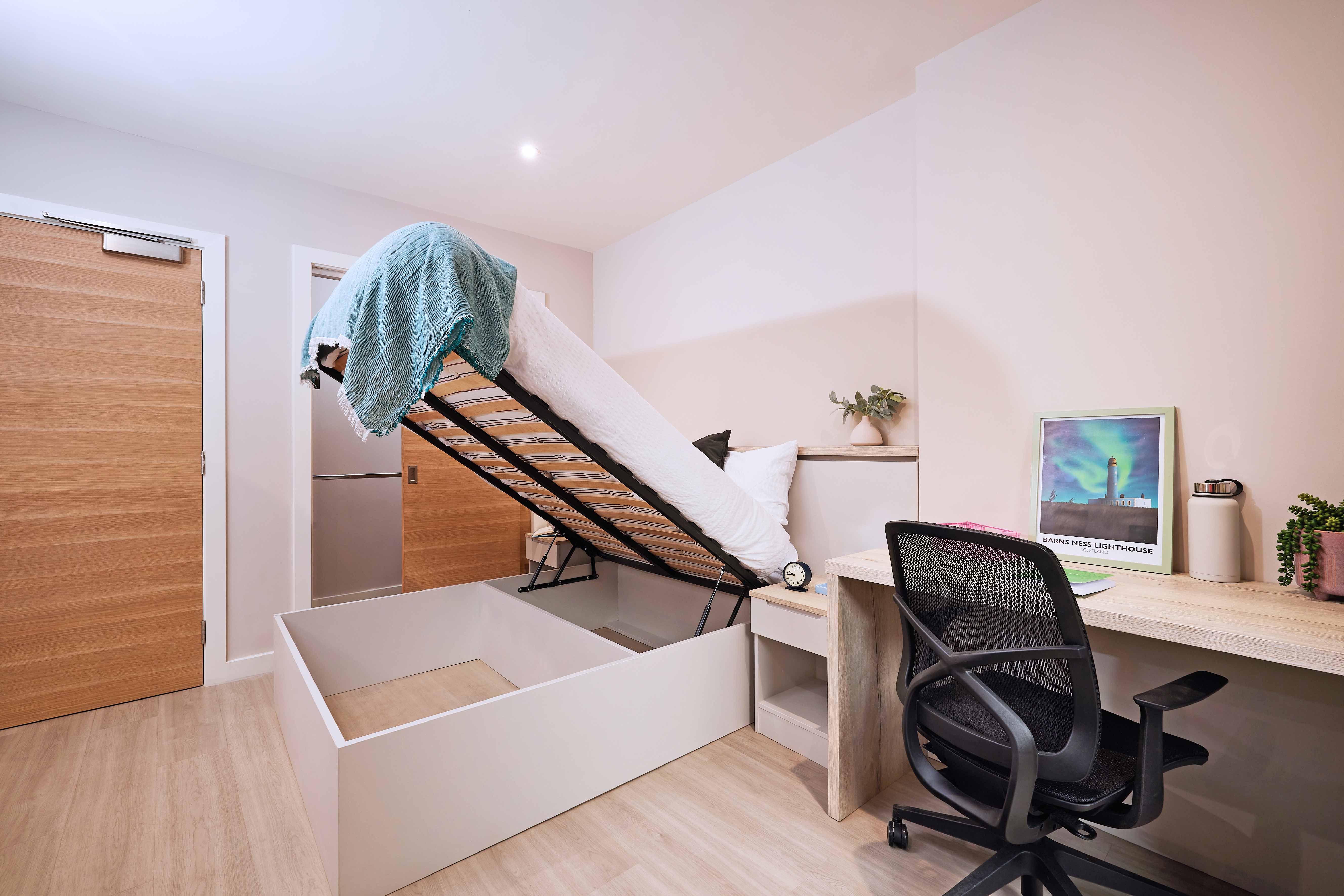 A modern bedroom with a bed lifted up for storage, revealing empty space underneath. A desk with a chair is on the right, featuring a framed picture, a laptop, a notebook, and a tumbler. The room has wooden floors and a door on the left wall.