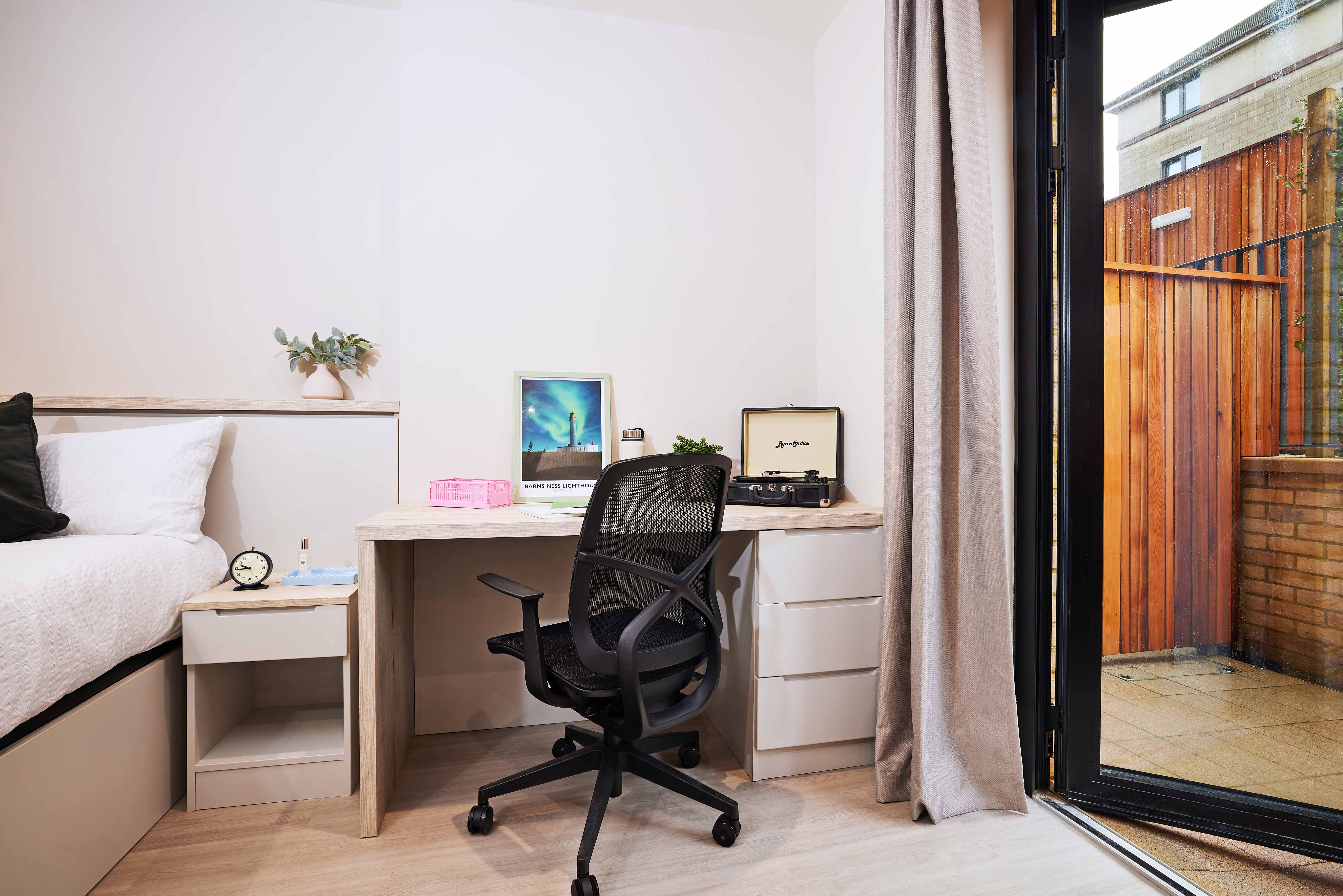 A modern bedroom features a bed, a desk with a black swivel chair, a framed abstract print, and an open record player. A sliding glass door leads to an outdoor space with wooden fencing. Neutral tones and minimalist decor create a cozy atmosphere.