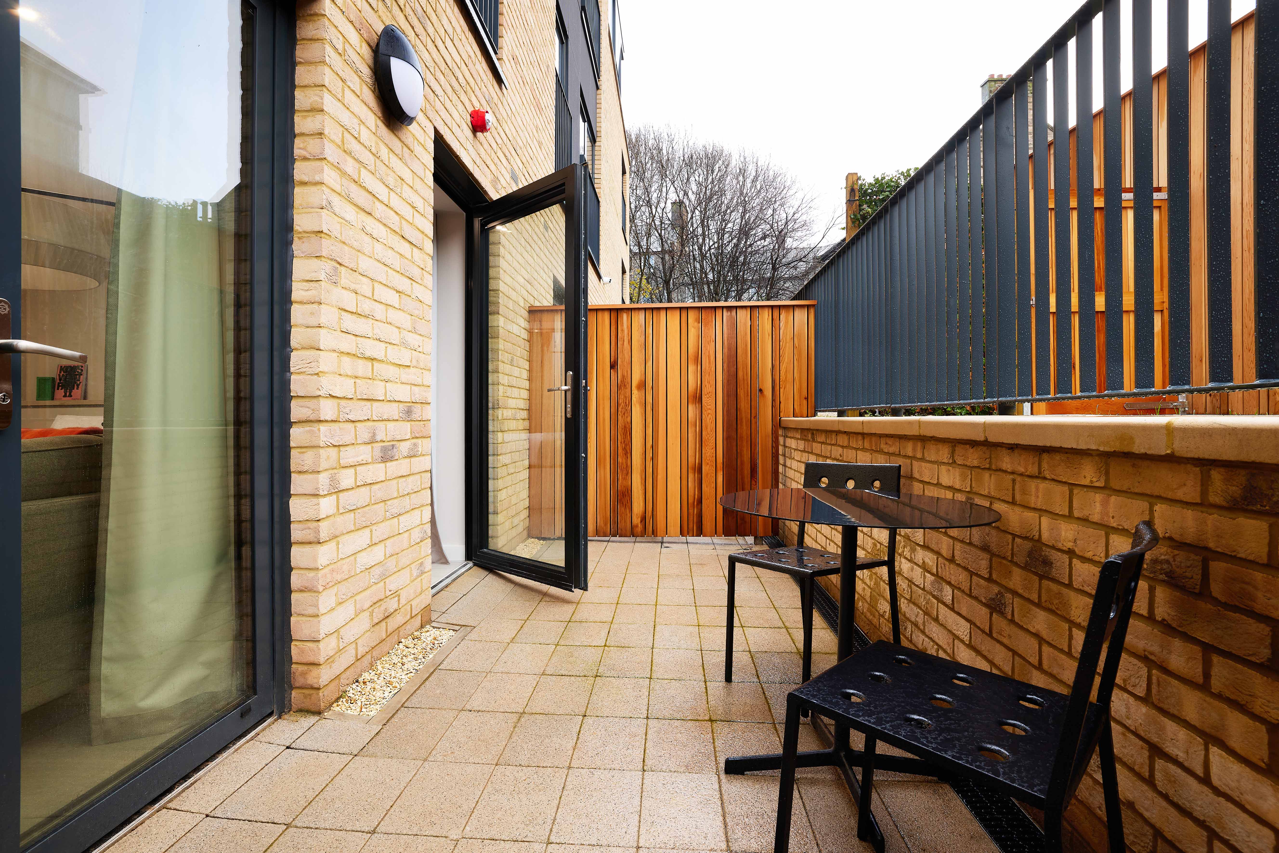 A small patio with two black metal chairs and a round table on a tiled surface. The area is enclosed by a wooden fence and brick wall, and a door with a glass panel leads indoors. Sparse trees are visible in the background.
