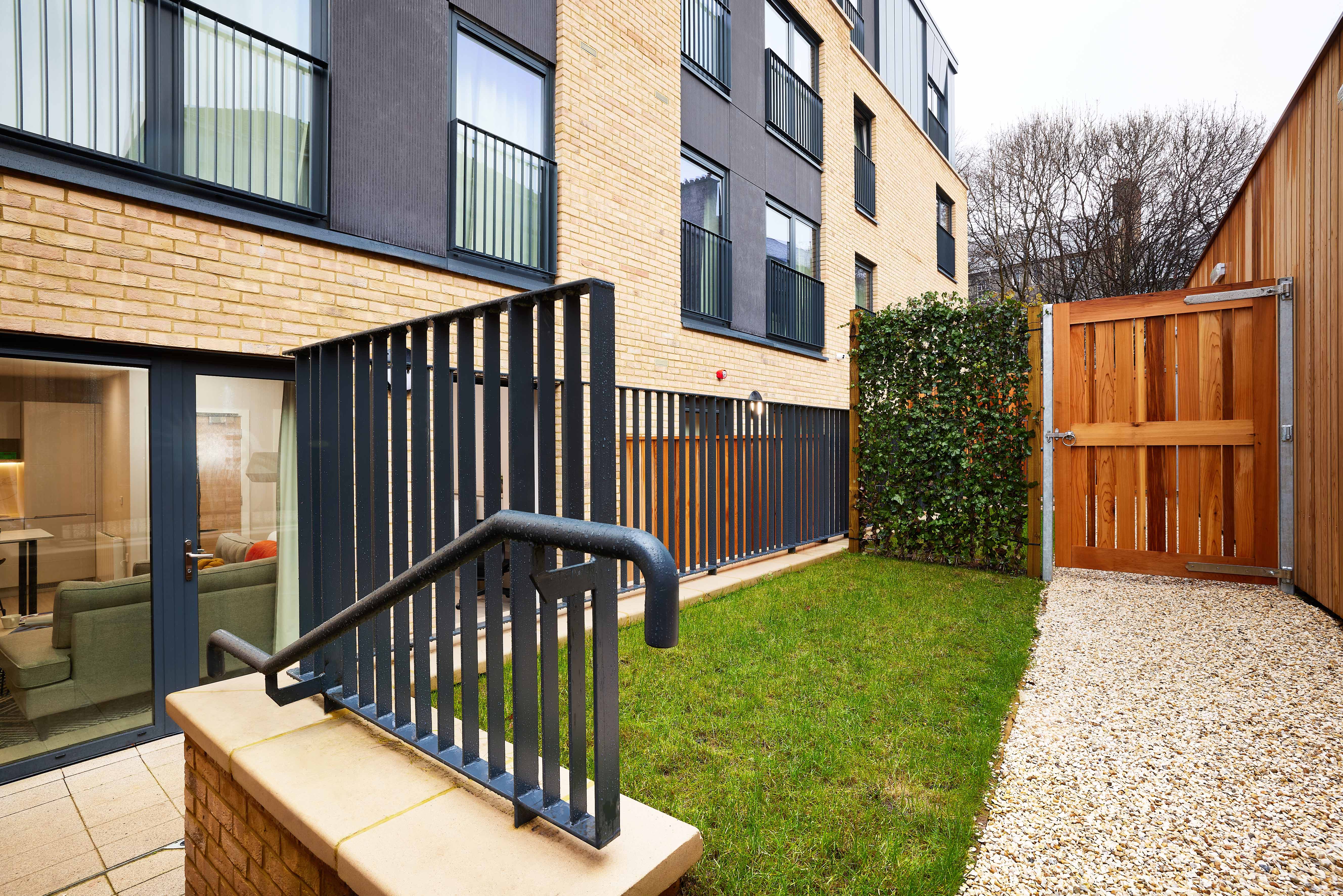 A small, enclosed outdoor space features a patch of green grass bordered by a gravel path leading to a wooden gate. Modern brick and dark exterior building with large windows in the background.