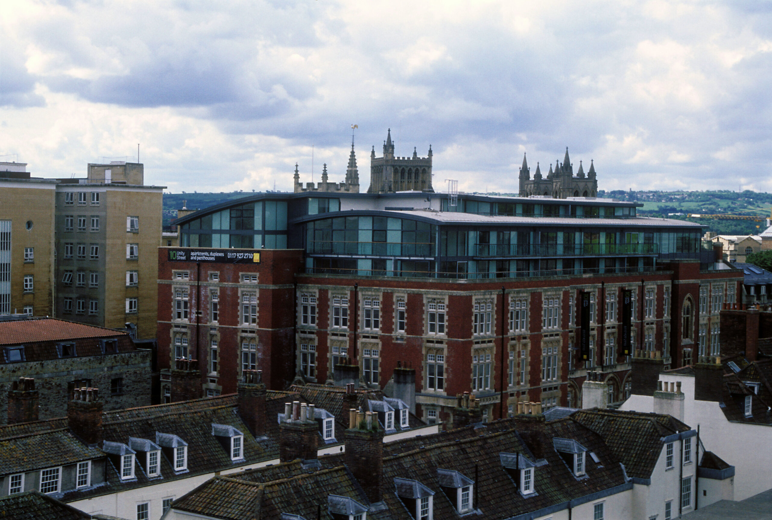 A cityscape featuring a modern rooftop extension on an older brick building. The structure stands amidst a mix of historic and contemporary architecture. In the background, church spires rise against a cloudy sky. Rooftops with chimneys are in the foreground.