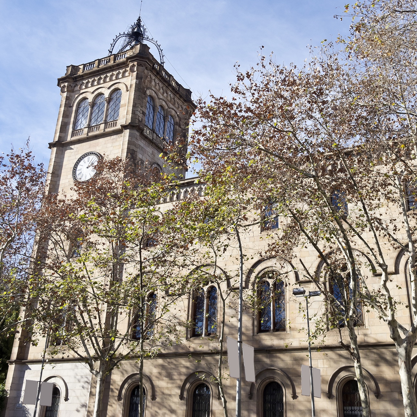 A historic building with a clock tower surrounded by bare trees in early spring or late autumn. The stone facade features arched windows, and the sky is clear and blue.