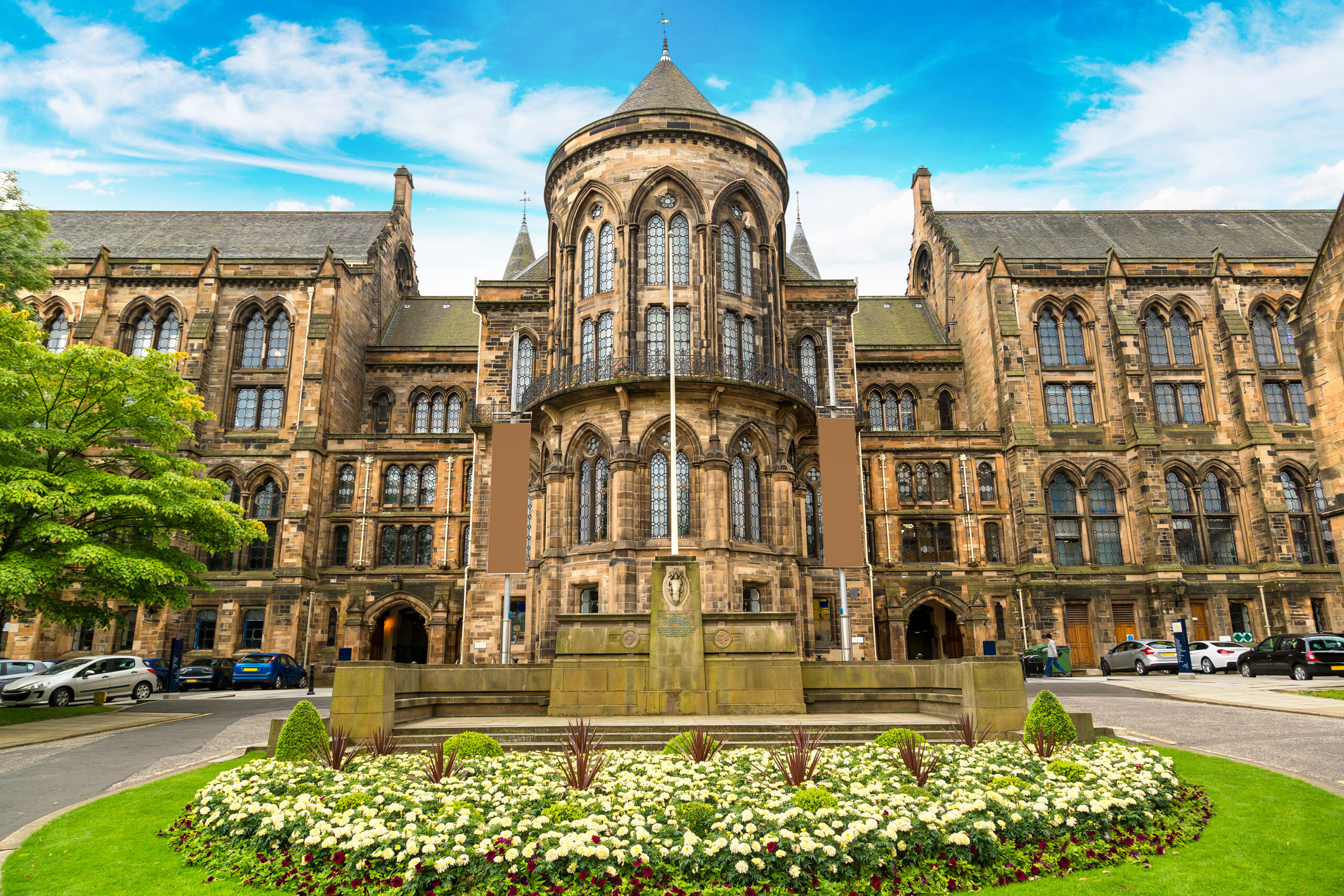 A historic, ornate stone building with a circular tower and spire stands against a blue sky. In the foreground, a circular garden bed is filled with white flowers and green shrubs. Parked cars line the driveway.