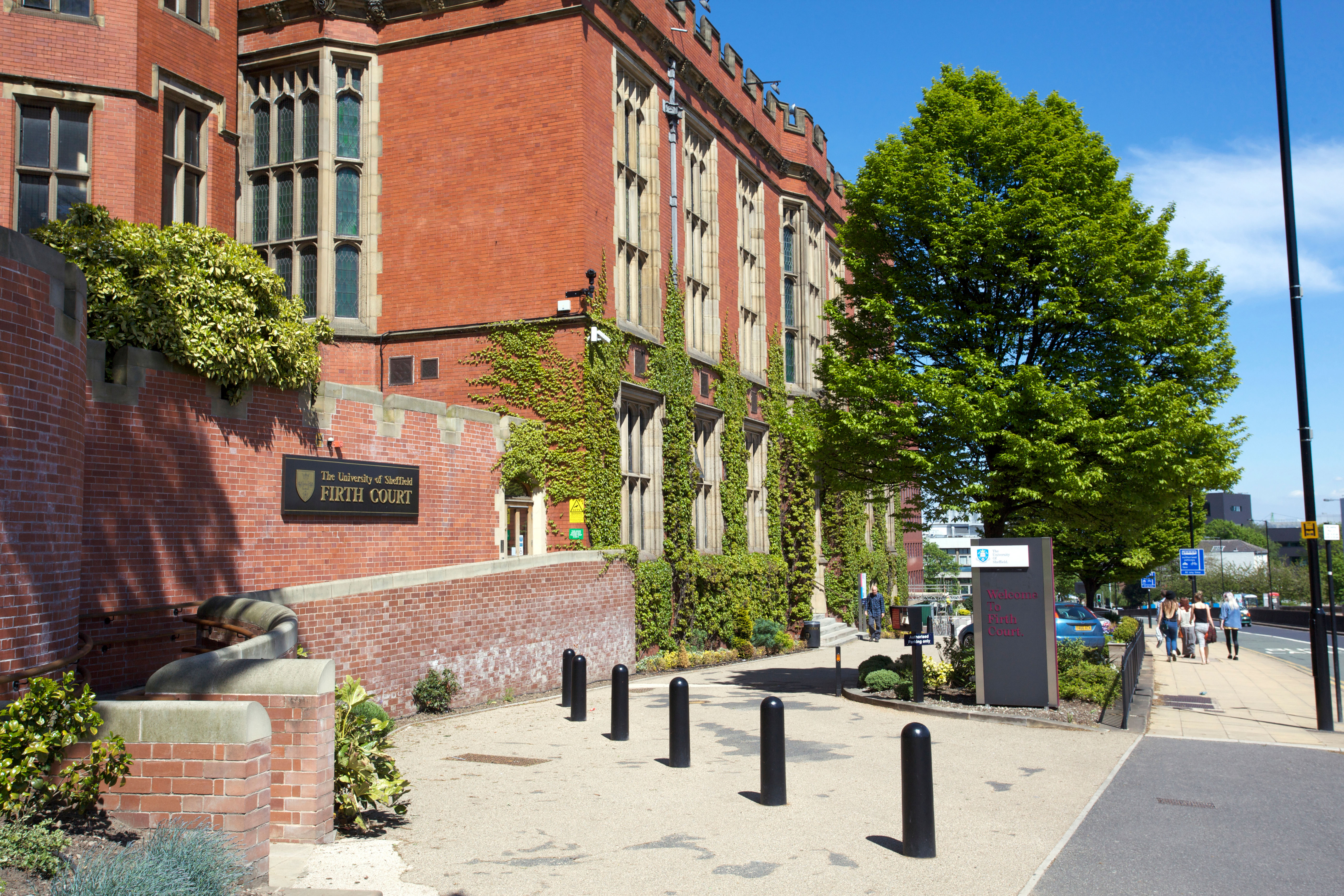 A large red-brick building with ivy-covered walls and tall windows, identified as Firth Court. A tree and signage are visible in the foreground. People are walking on the adjacent sidewalk under a clear blue sky.