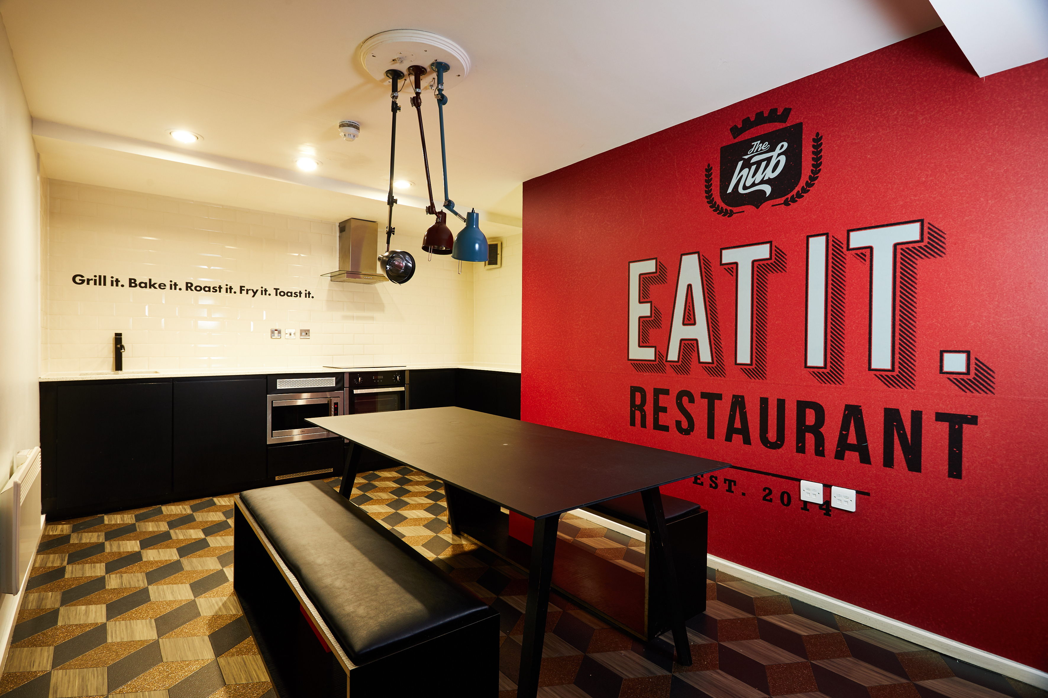 A modern kitchen with a black table and stools in the center. The red wall displays the logo "Hub: Eat It. Restaurant" with a slogan. The opposite white tiled wall mentions cooking methods. Industrial-style lighting hangs from the ceiling.