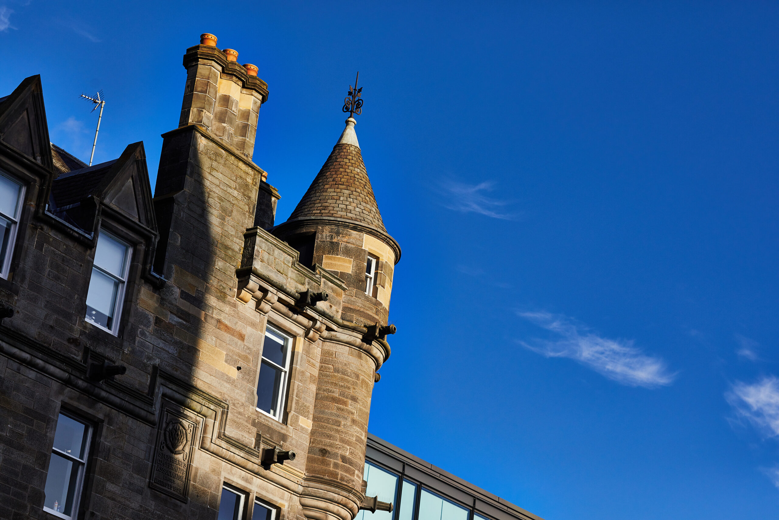 A stone building with a small turret and chimney, featuring decorative architecture under a clear blue sky. The building's details stand out sharply in the bright sunlight.