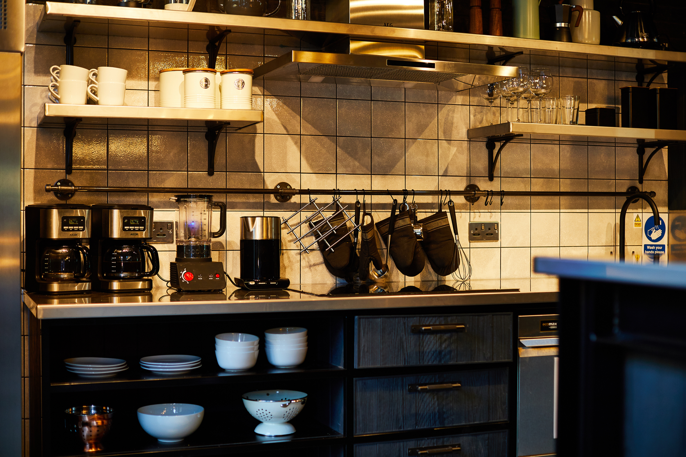 A modern kitchen in Fountainbridge with a tiled backsplash features two coffee makers, a blender, and a pot on the countertop. Shelves above hold cups, glasses, and dishes. Below are drawers and a shelf with bowls and a colander. Oven mitts hang near outlets.