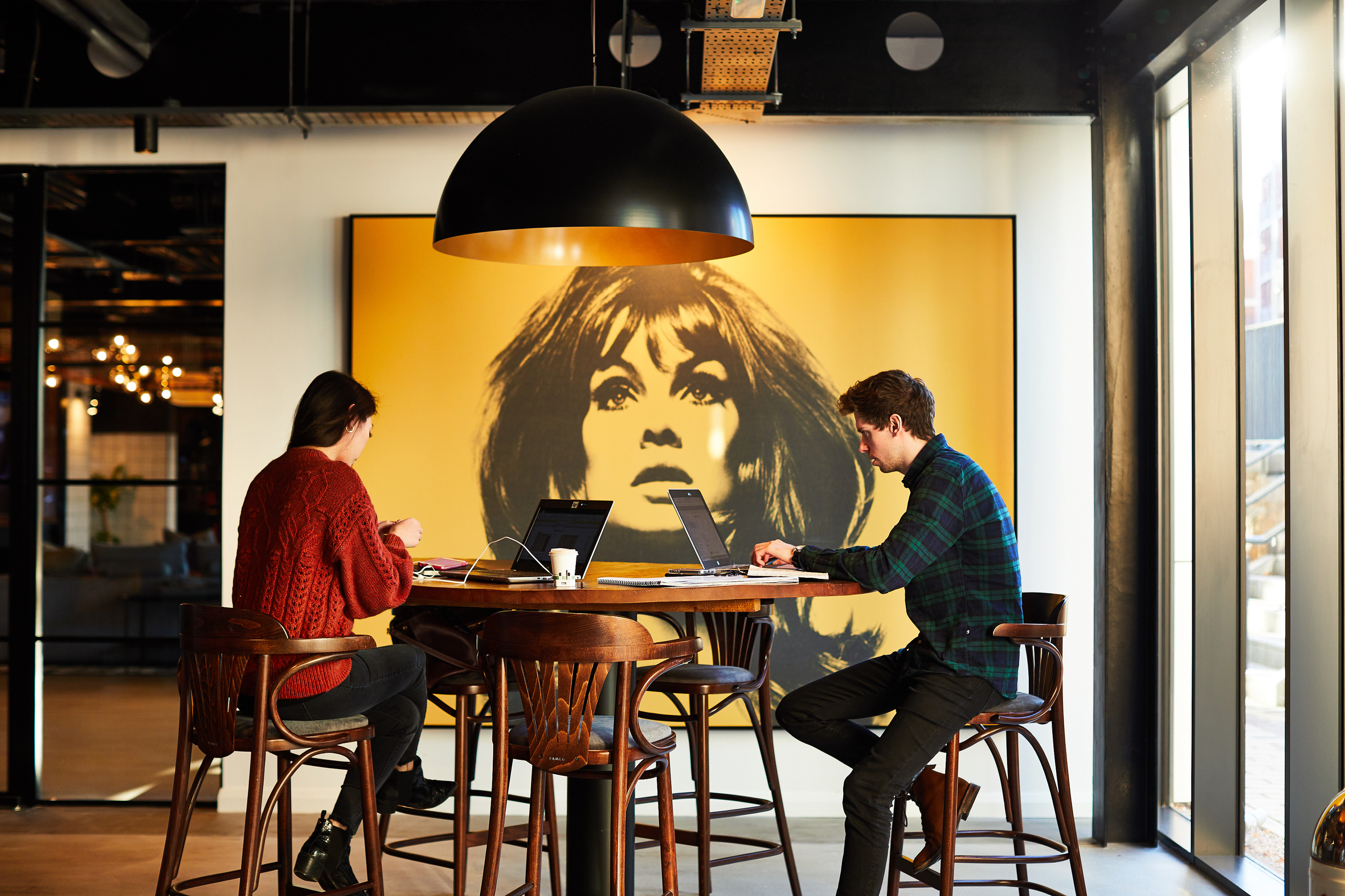 Two people work on laptops at a wooden table in a modern Fountainbridge workspace. Behind them is a large yellow portrait of a woman's face. The room is well-lit with a black overhead light and expansive windows that let in plenty of natural light.