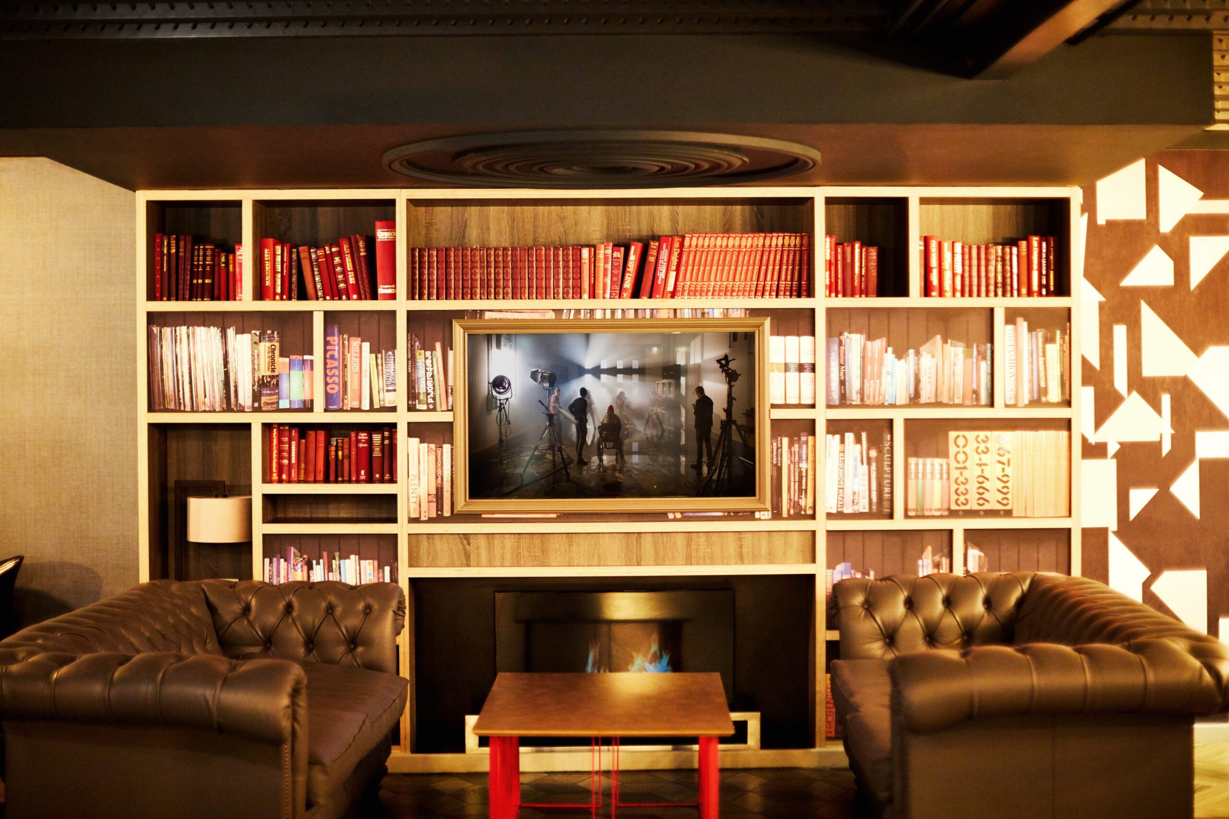 A cozy library setting with two brown leather sofas facing each other. A large bookshelf filled with red and white books surrounds a TV playing a black-and-white scene. A small wooden coffee table sits between the sofas.
