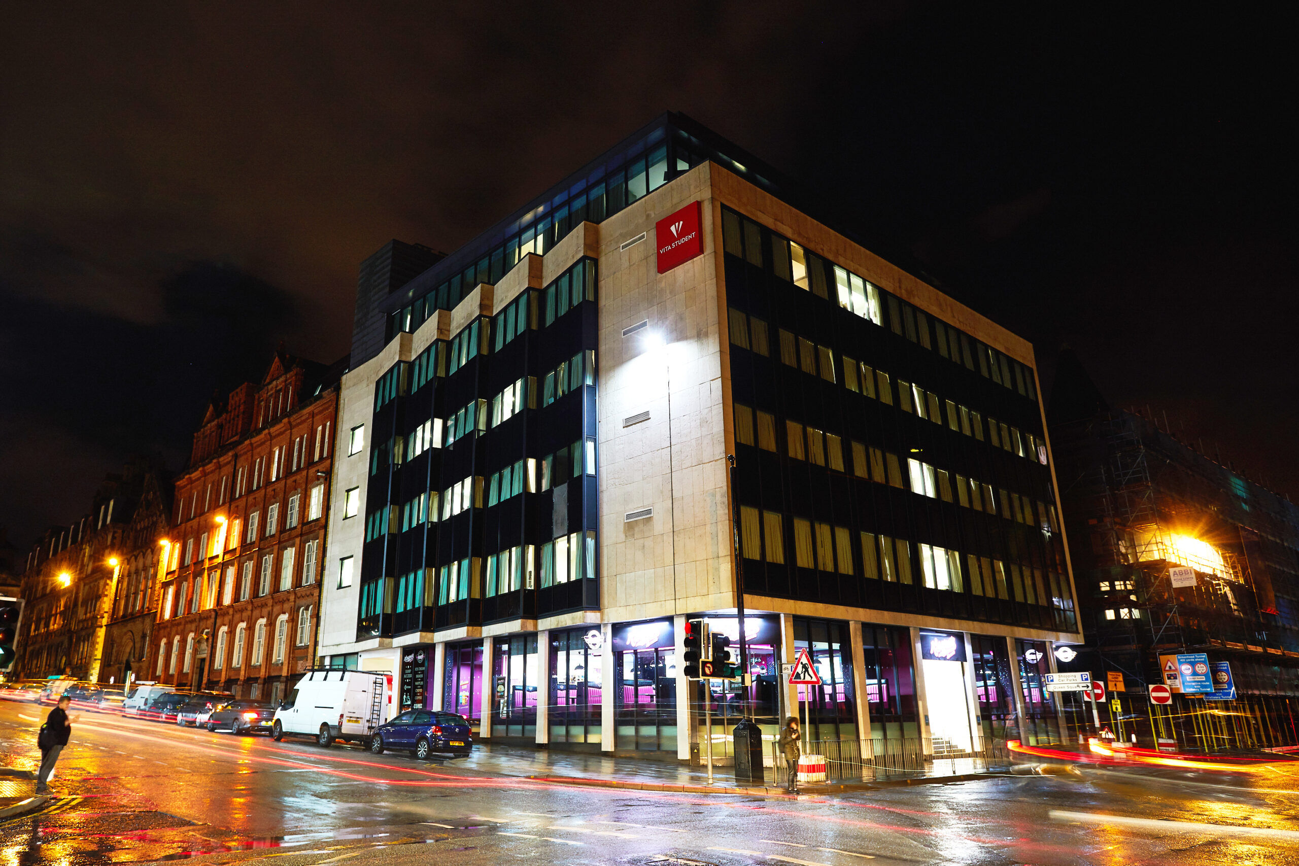 A modern, multi-story building with a red logo near the top corner is lit up against the night sky. The streets are wet and reflective, with light trails from passing vehicles. Nearby, a historic brick building adds contrast.