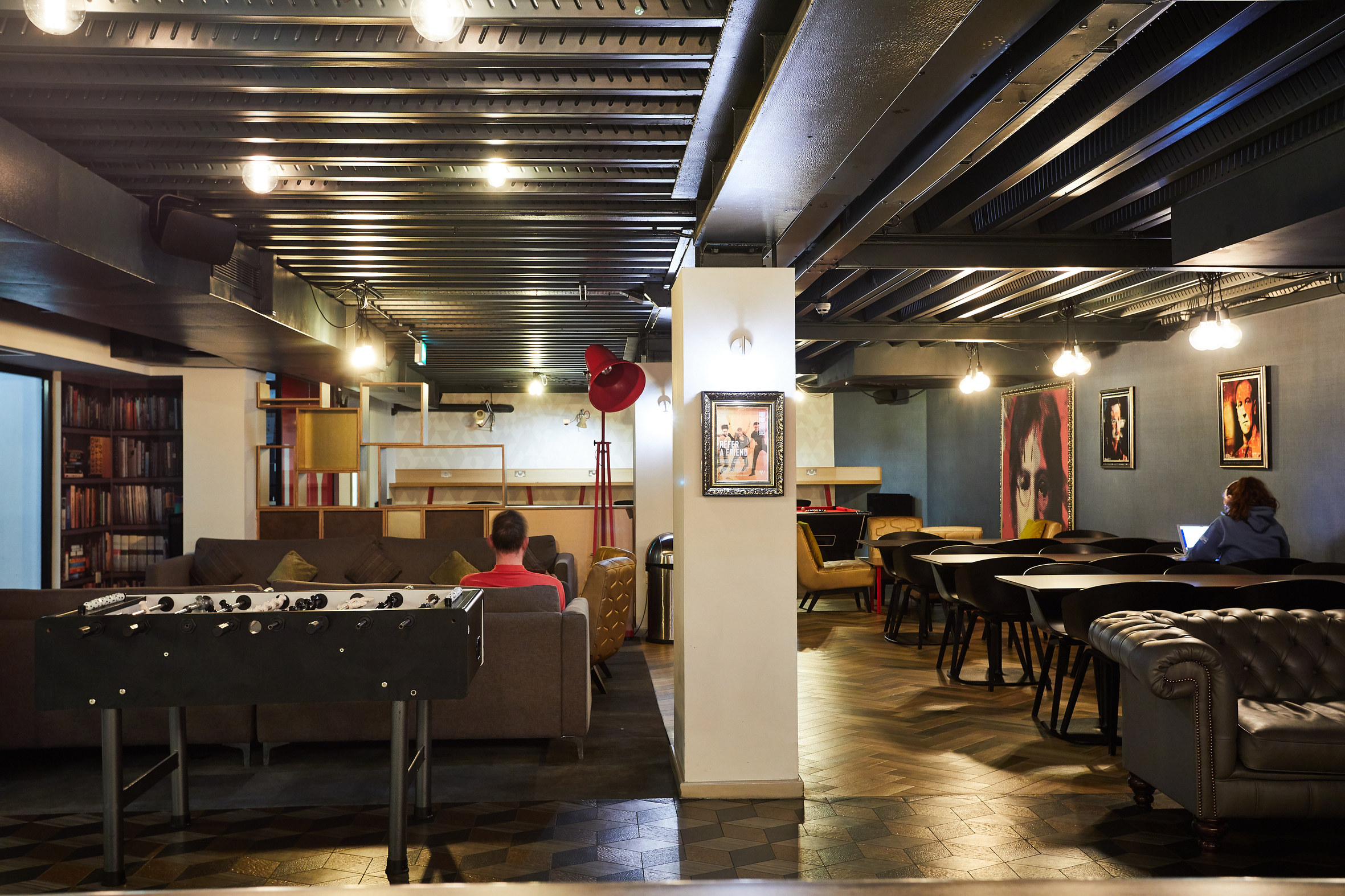 A modern lounge area in luxury student accommodation features sofas, a foosball table, and vibrant artwork on the walls. Two people are seated, one using a laptop. Shelves brimming with books add charm to the space, which is illuminated by sleek overhead lights.
