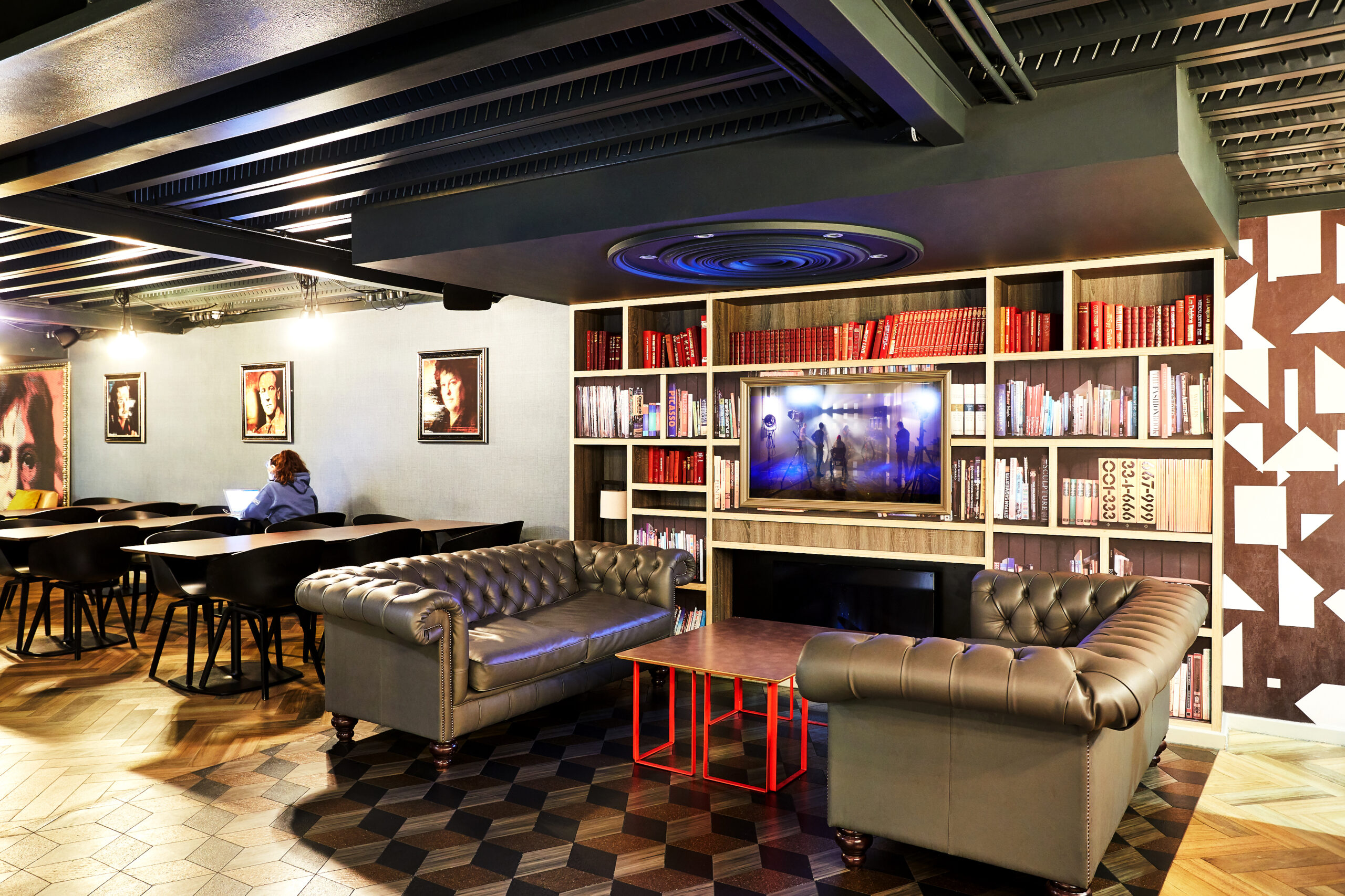 A cozy room features leather sofas, a wooden coffee table, and a large bookshelf with red and white books. A TV displays an image among the shelves. Portraits adorn the walls, and a person sits at a dining table in the background.