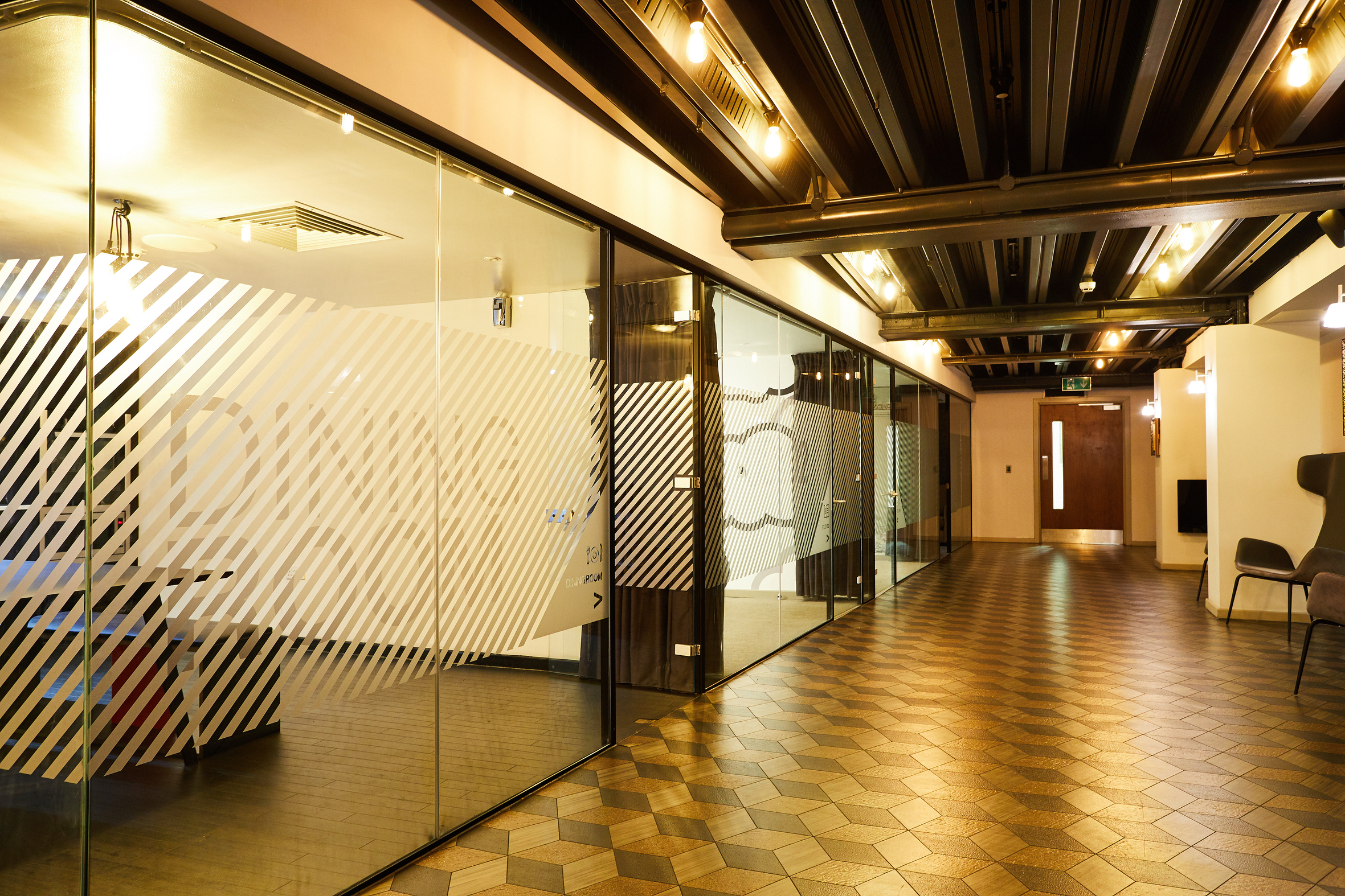 The modern office hallway evokes the elegance of luxury student accommodation, featuring glass-walled meeting rooms with frosted diagonal stripes on the left. The hexagonal tile floor and warm lighting from exposed ceiling bulbs complete the sophisticated ambiance.