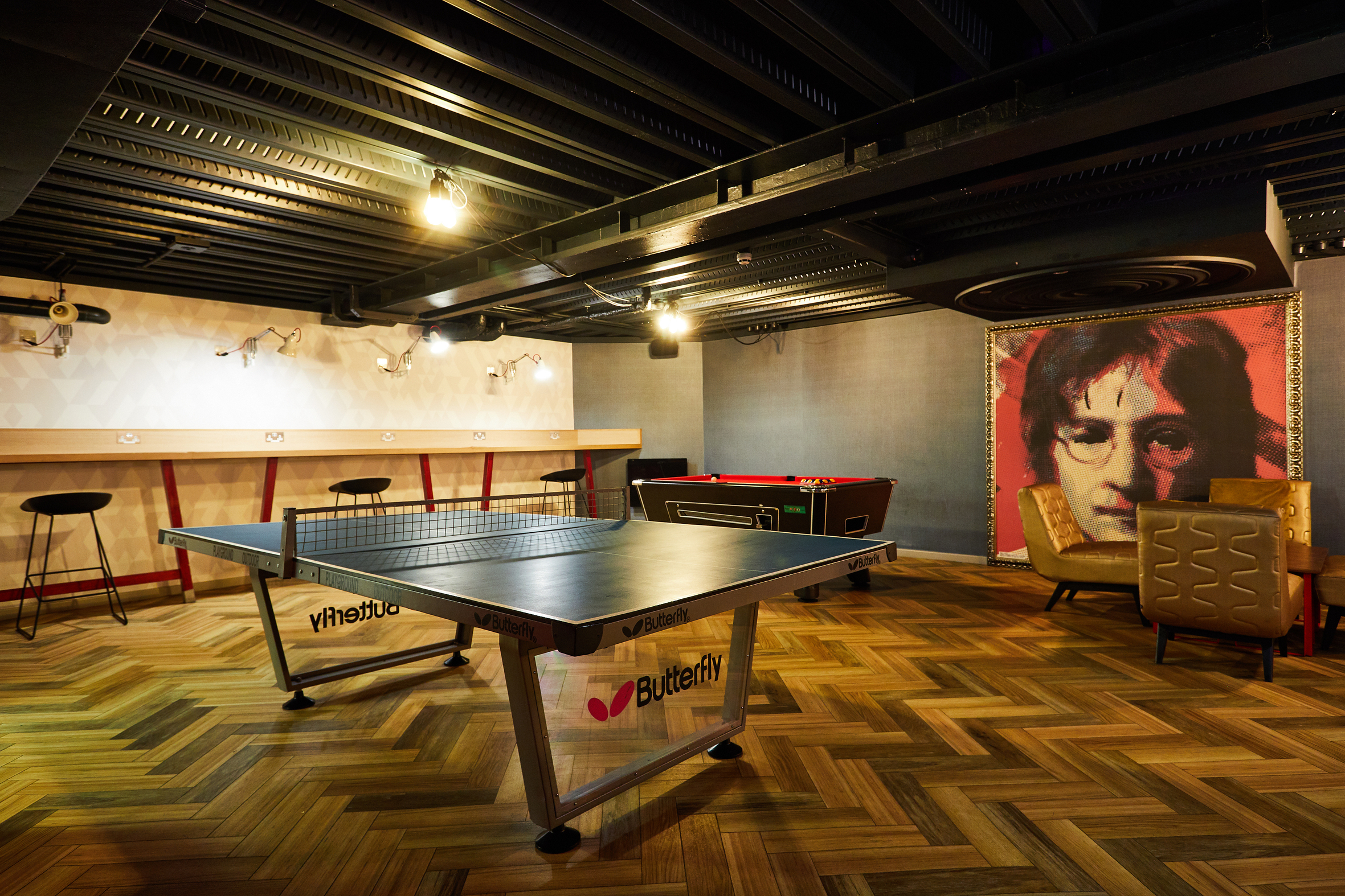 The luxury student accommodation boasts a vibrant room with a ping pong table, foosball table, bar stools, and a large portrait on the wall. The floor showcases patterned wood, while the ceiling is adorned with exposed beams and lights for a chic touch.