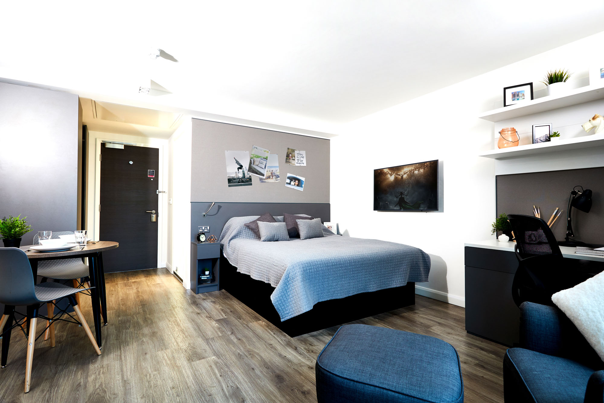 A modern apartment with a double bed, gray bedding, and pictures on the wall behind. The square wall-mounted TV faces a desk with a chair and shelves. A small dining table with chairs and wooden flooring complete the cozy space.