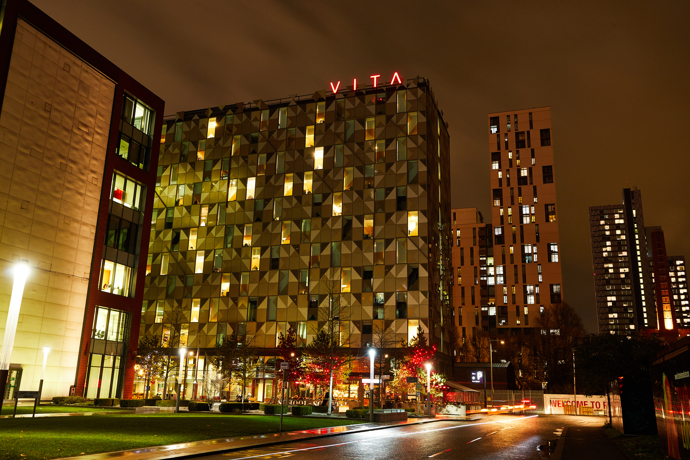 Night view of a modern building with the sign "VITA" on top, featuring a geometric facade on First Street. The scene is illuminated by streetlights and glowing windows, with traffic lights streaking on the road in the foreground and high-rise buildings in the background.