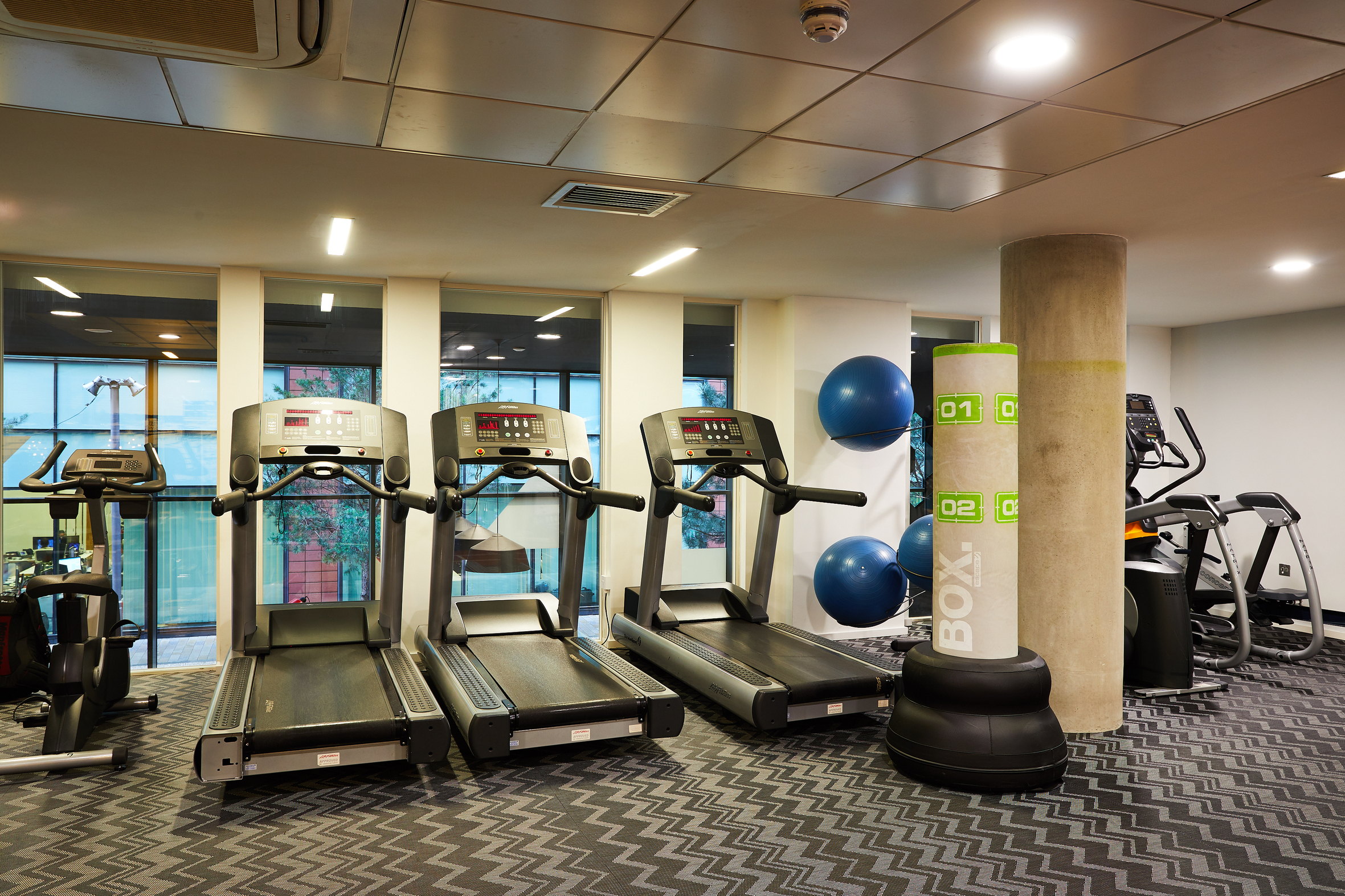 Nestled on First Street, this modern gym boasts three treadmills, exercise bikes, a weighted punching bag, and stability balls. Large windows offer a view of the trees outside, while zigzag-patterned flooring and overhead lighting enhance the welcoming space.