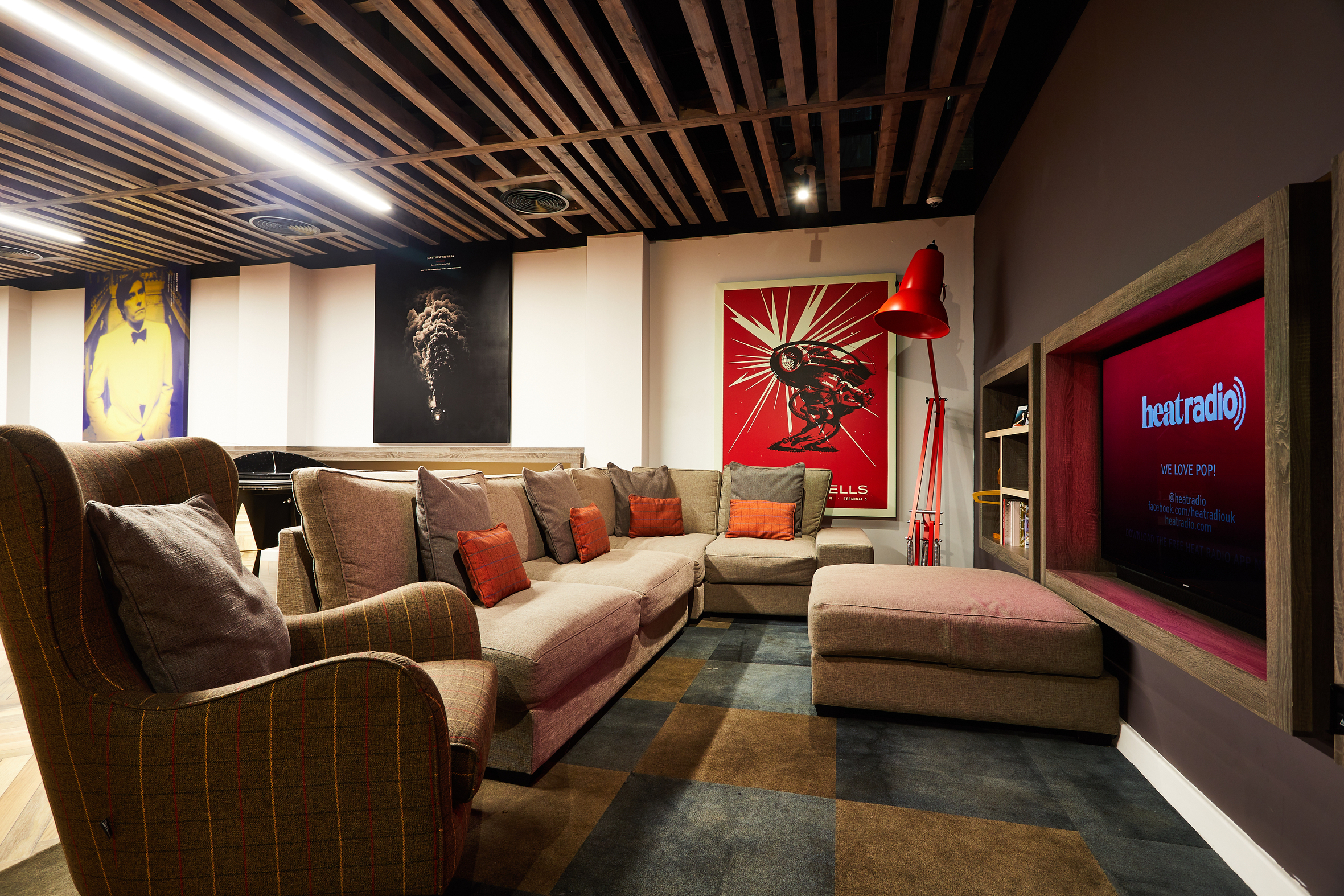 A cozy lounge space with a sectional sofa and armchairs, featuring red cushions and a red floor lamp. The wall displays colorful art posters. A TV screen shows a radio station logo. The ceiling showcases exposed wooden beams.