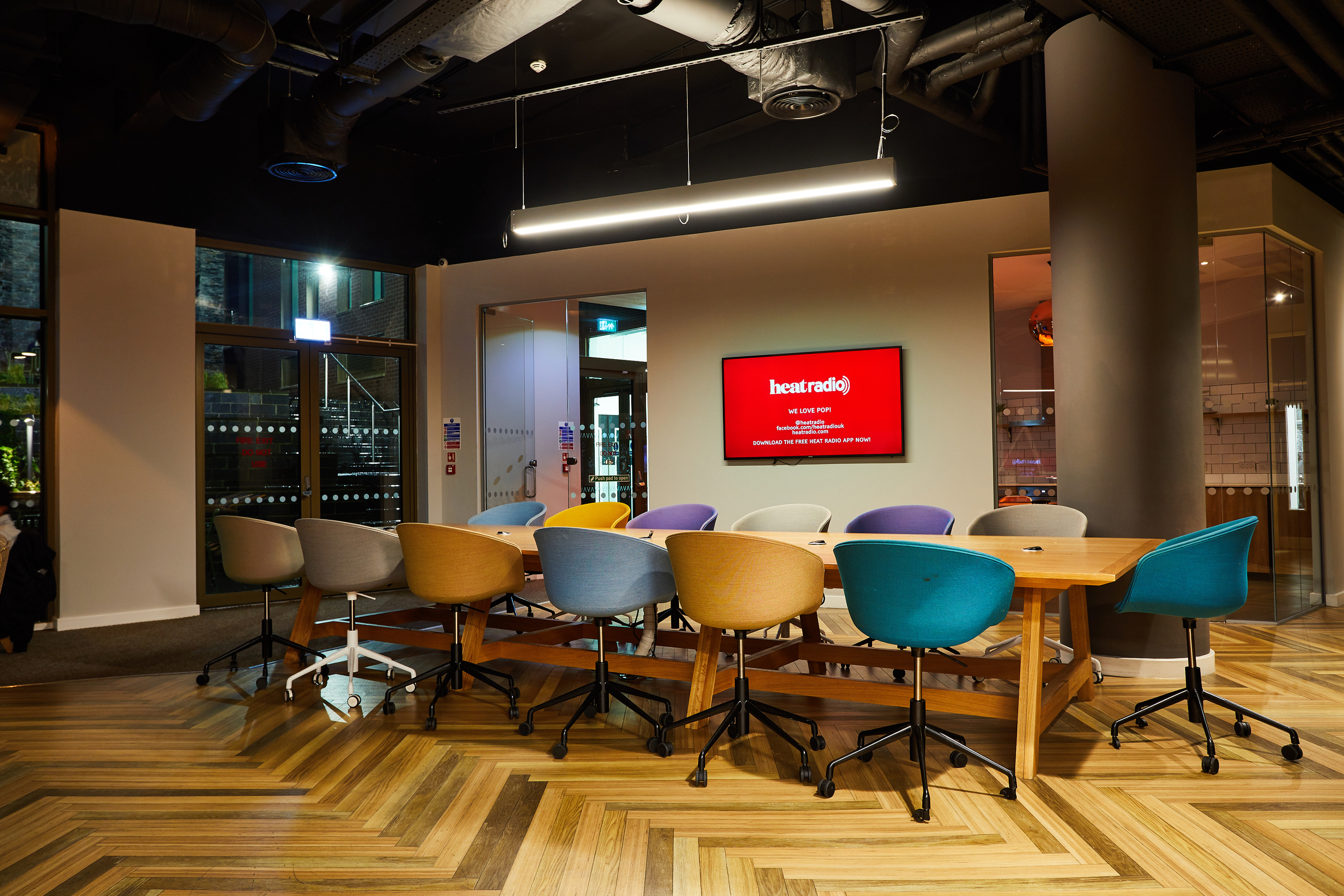 A modern meeting room with a long wooden table surrounded by colorful office chairs. A red screen on the back wall displays the "Heart Radio" logo. The room features wooden flooring and a ceiling with exposed fixtures.