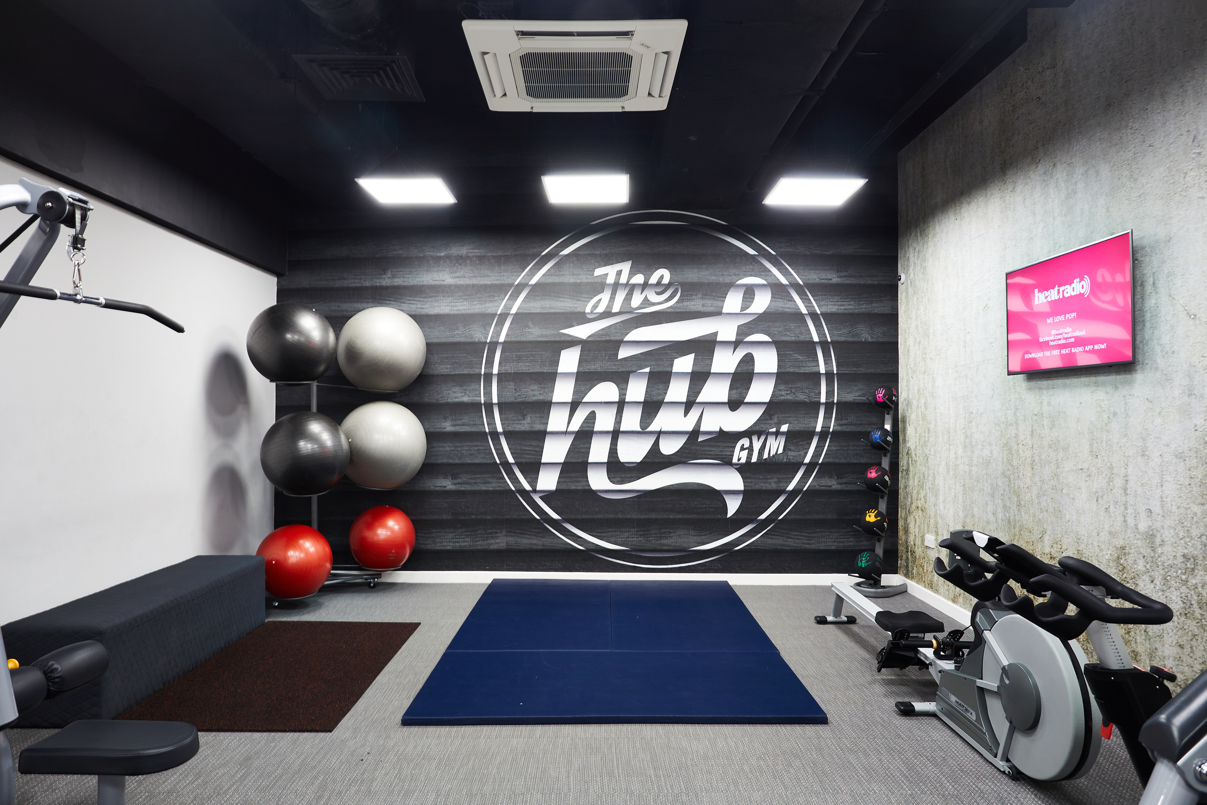 A modern gym room with exercise equipment including a stationary bike, workout benches, and exercise balls. The walls have "The Hub Gym" logo and a small pink digital screen displaying information. The room is well-lit with ceiling lights.