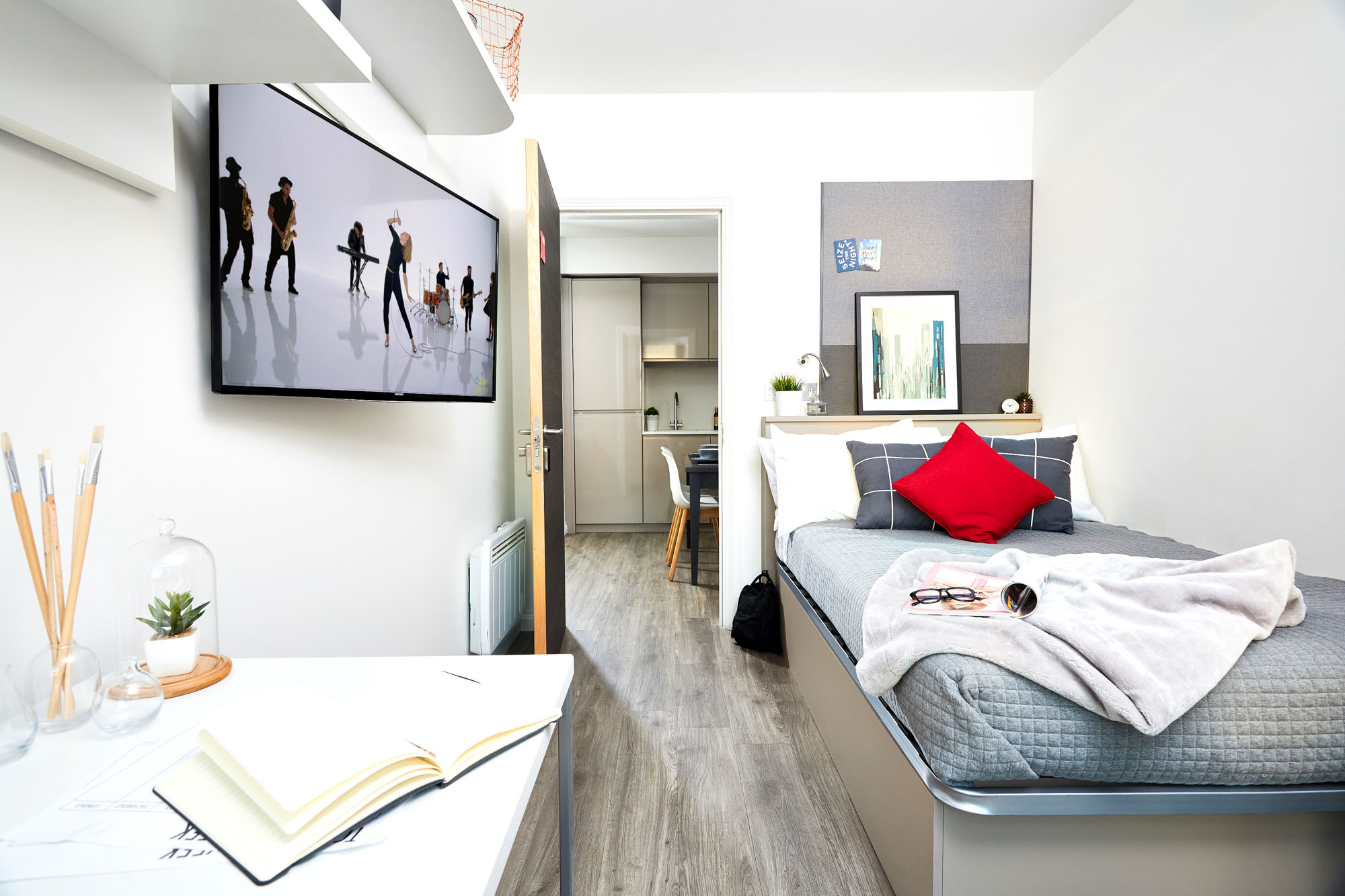 A modern, bright dorm room in Telephone House features a bed on the right, adorned with gray bedding, red and gray pillows, and a fluffy throw. A flat-screen TV showing dancers is mounted on the wall. There's a desk with an open book, glasses, and a cozy nook in this stylish real estate offering.
