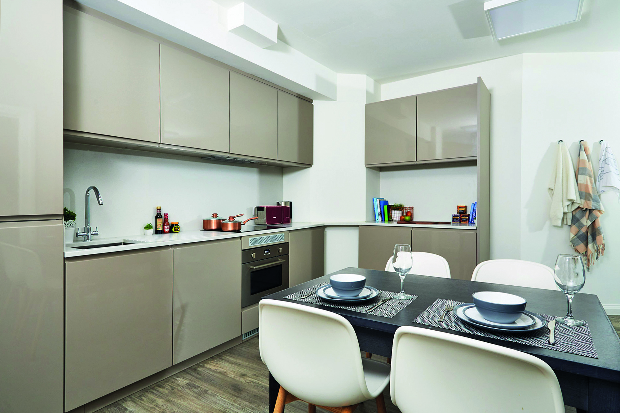 Modern kitchen with beige cabinets and a sleek black dining table set for three. The space features an oven, cooktop with copper pots, and various condiments. Towels hang on the right wall next to a vintage telephone, and books are neatly placed on top of corner cabinets.