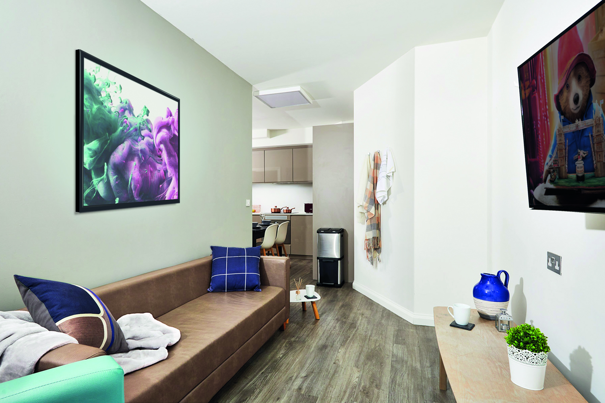 The modern living room in Telephone House features a wooden floor, brown couch, and pillows. A colorful abstract painting adorns the wall. A TV on the right displays a movie scene. In the foreground, a small table with a plant and blue pitcher sits, offering views into the spacious kitchen beyond.