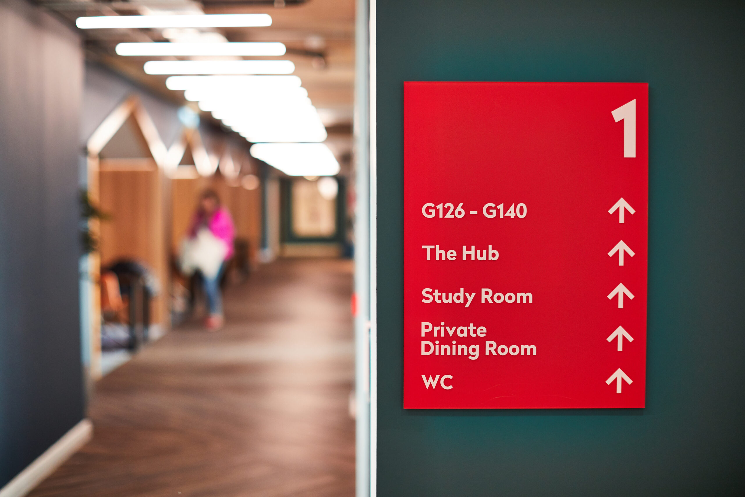 A hallway with a bright red sign displaying arrows and room names: "G126-G140," "The Hub," "Study Room," "Private Dining Room," and "WC." A person is walking in the blurred background.