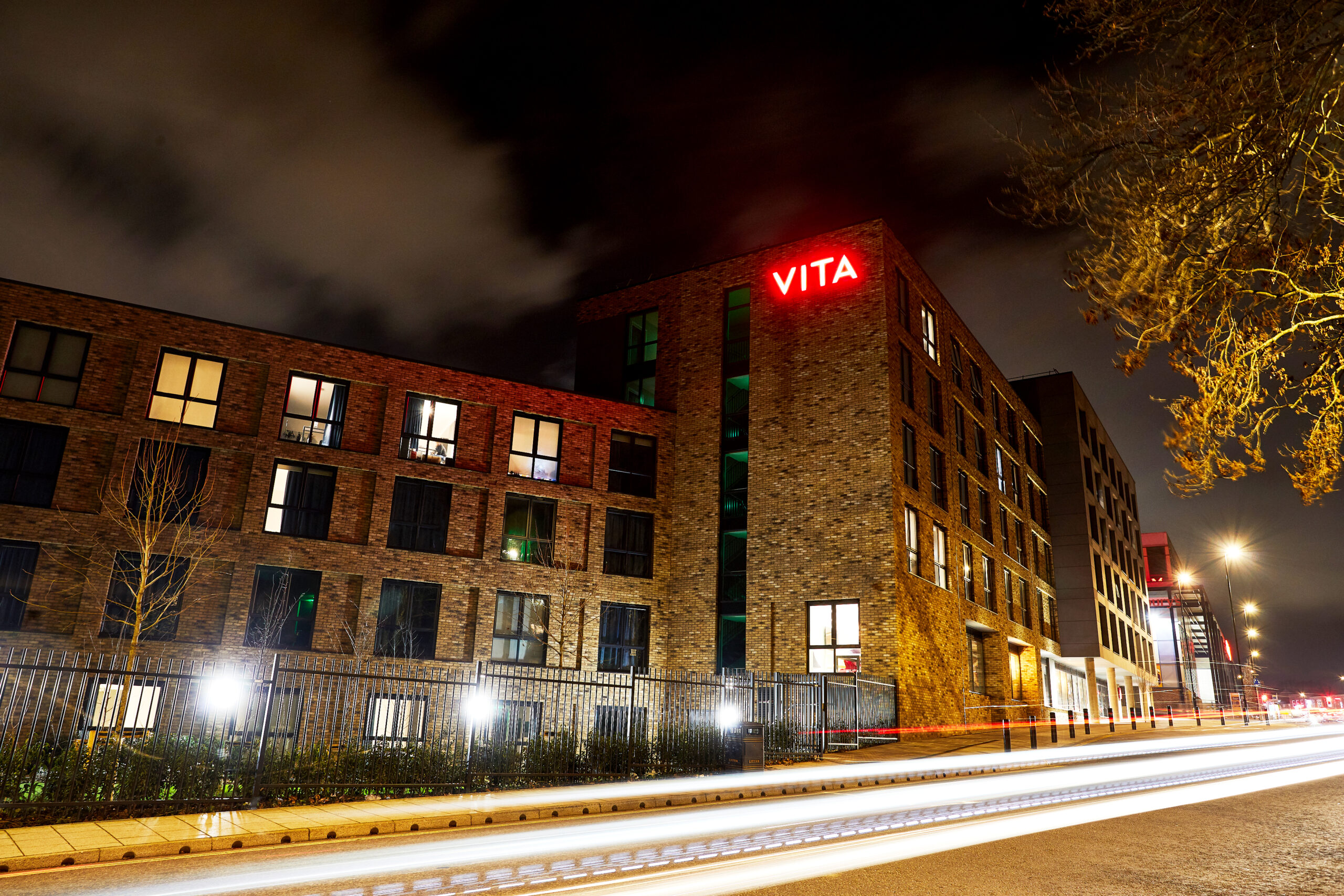 Exterior of a modern brick building with "VITA" in red neon on the top. The sky is cloudy, and trees are in the foreground. The street below shows light trails from passing vehicles, indicating night-time traffic.