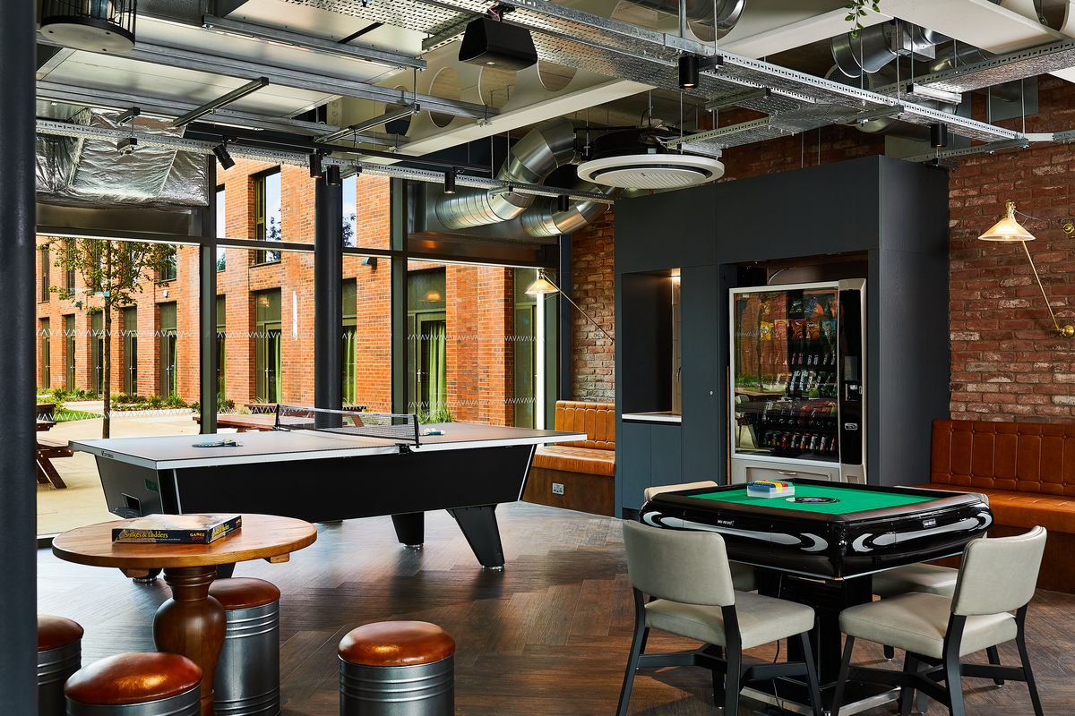 Modern recreational room with a pool table, round wooden table, stools, and a card table with chairs. Exposed brick walls and ceiling ductwork create an industrial feel. Large windows offer a view of a brick building and greenery outside.