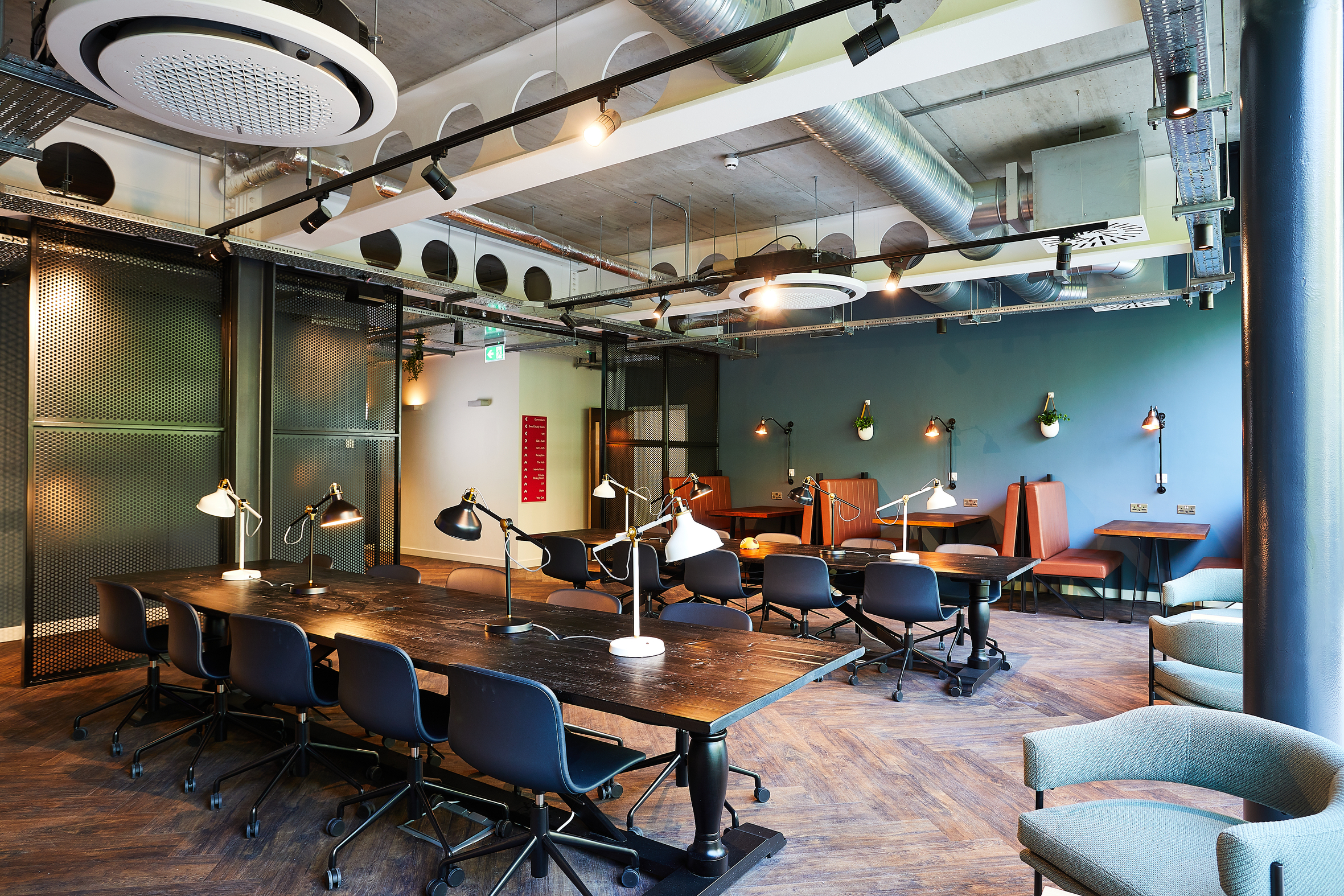 A modern coworking space with dark wooden tables, black chairs, and retro desk lamps. The room features exposed ductwork and overhead lighting. Teal walls and brown seating booths are in the background, creating a stylish industrial atmosphere.
