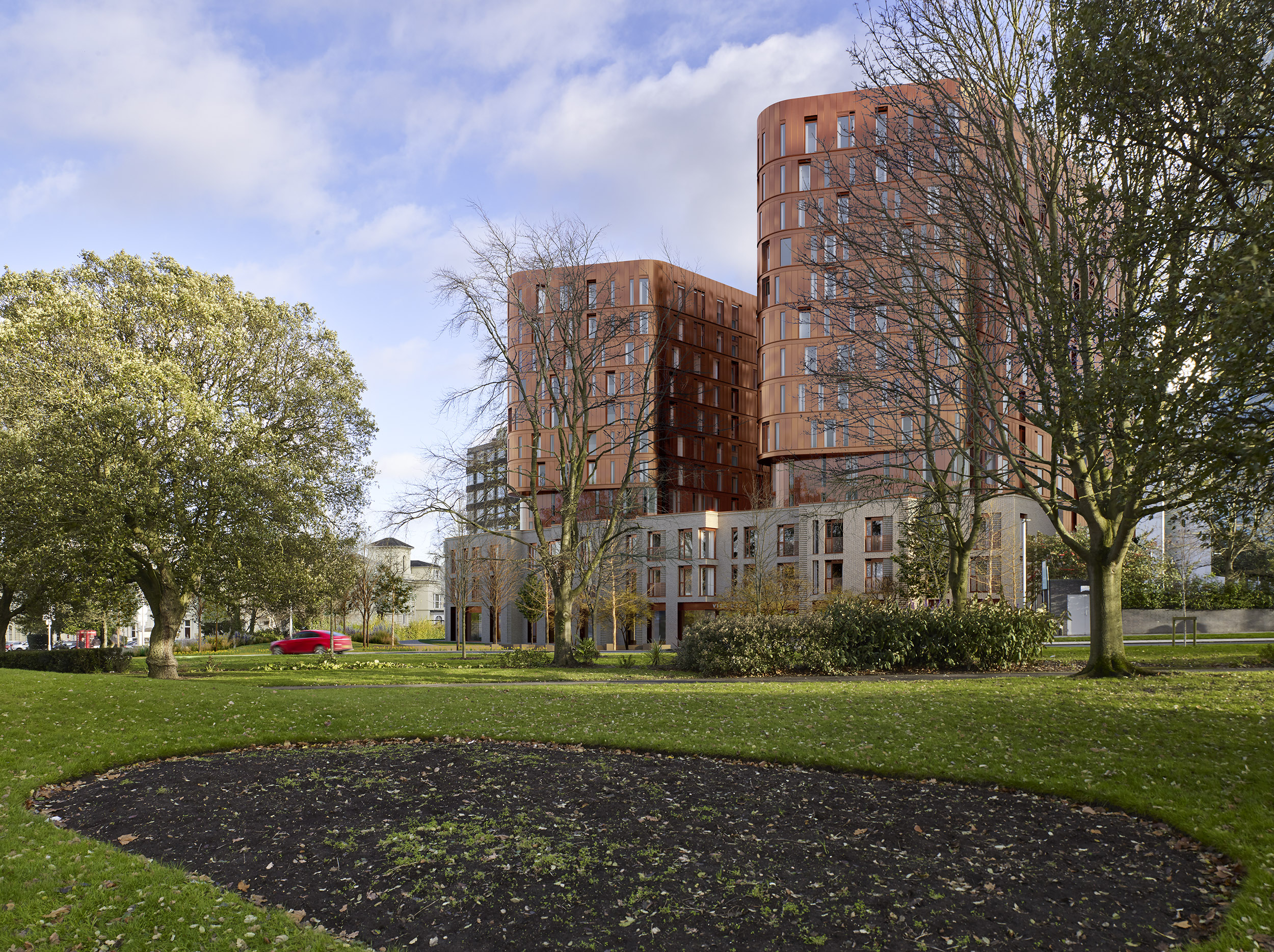 A modern, cylindrical brick building with large windows is nestled among trees and grass in a park setting, serving as a lively student accommodation in Coventry. The sky is partly cloudy, and there's a red car visible in the distance.