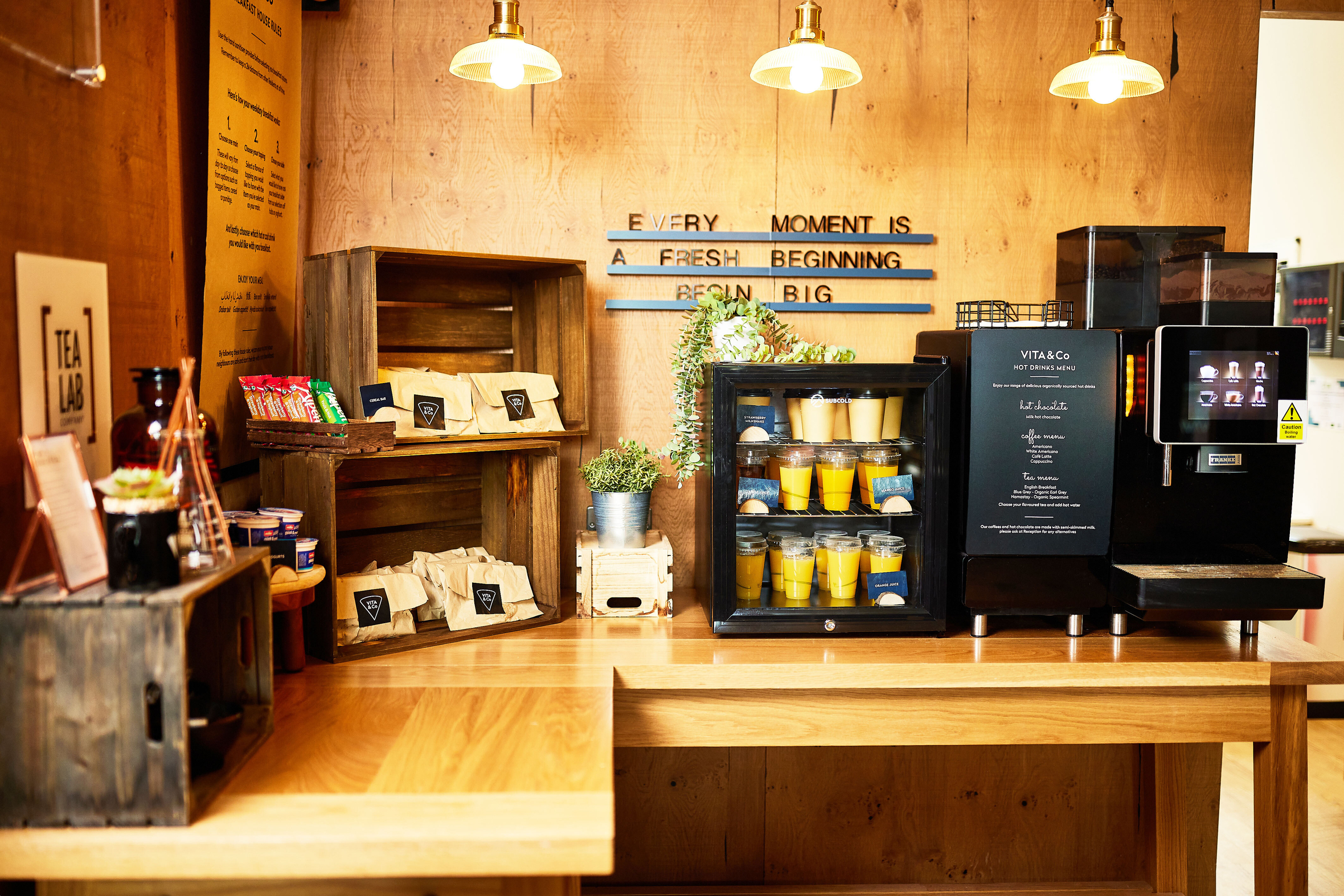A cozy cafe setup with a wooden counter displaying assorted tea bags and a variety of juices in a mini-fridge creates an inviting circle of warmth. A coffee machine hums nearby, while plants and a wall sign reading "Every moment is a fresh beginning. Begin big." add a welcoming touch.