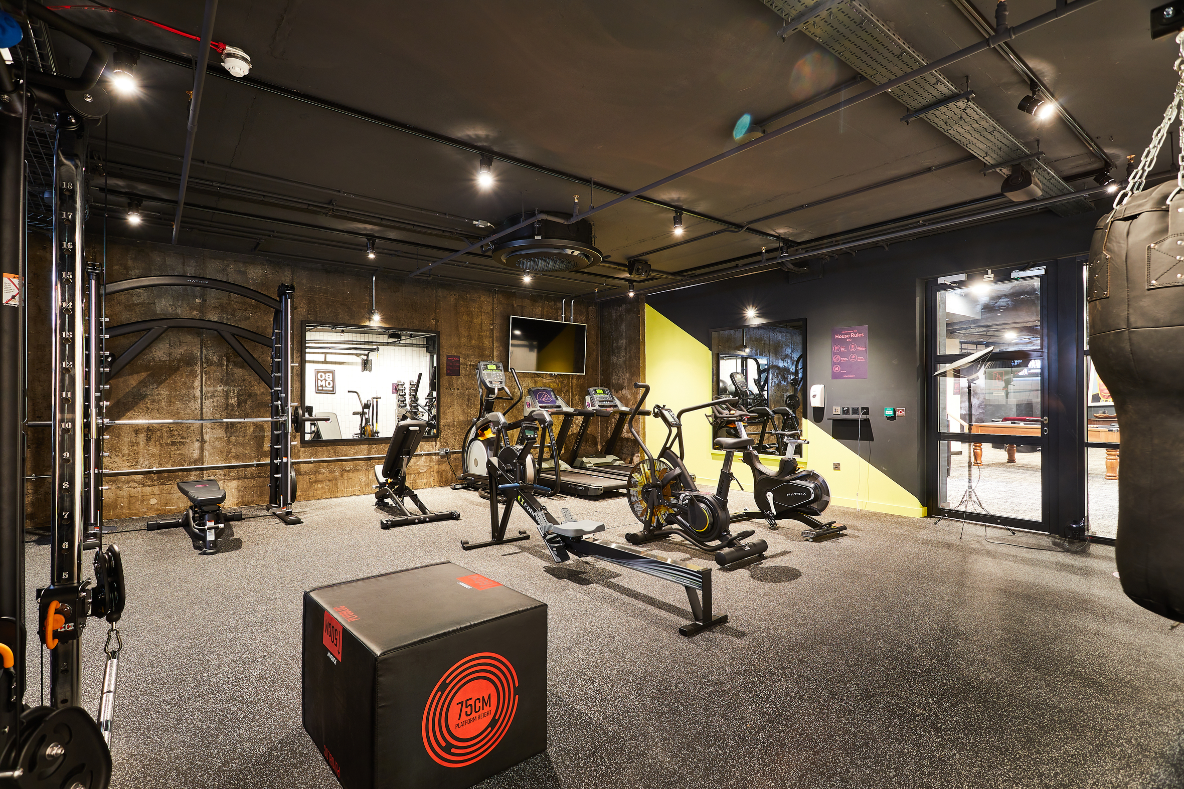 A modern gym on Portland Crescent features an array of exercise equipment, including stationary bikes, a rowing machine, and a boxing area with a punching bag. The dark and bright yellow walls contrast with the squat rack and soft plyometric box in the foreground.