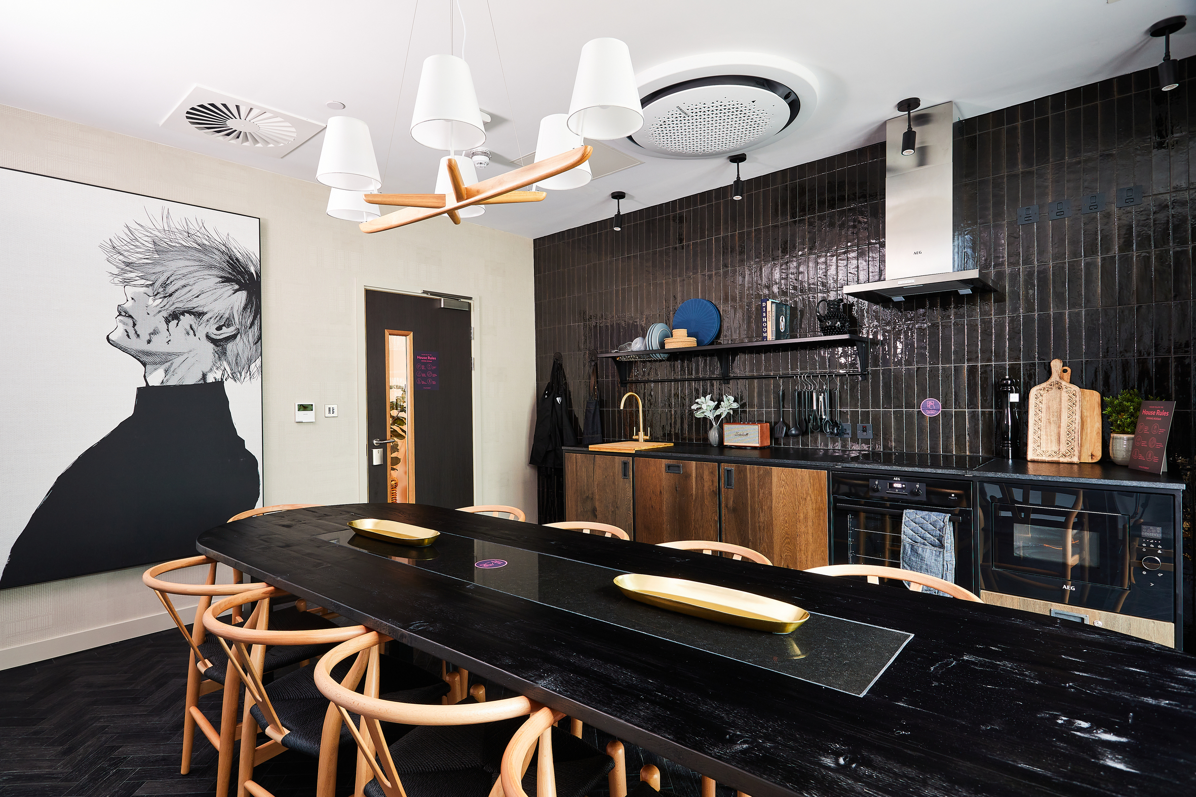 Located in the heart of Portland Crescent, this modern kitchen features black tiled walls and wooden cabinetry. A long black dining table with chairs stands at the center, while a large black and white portrait adorns the wall. Overhead, white ceiling lights and a circular vent offer a sleek touch amidst decorative shelves.