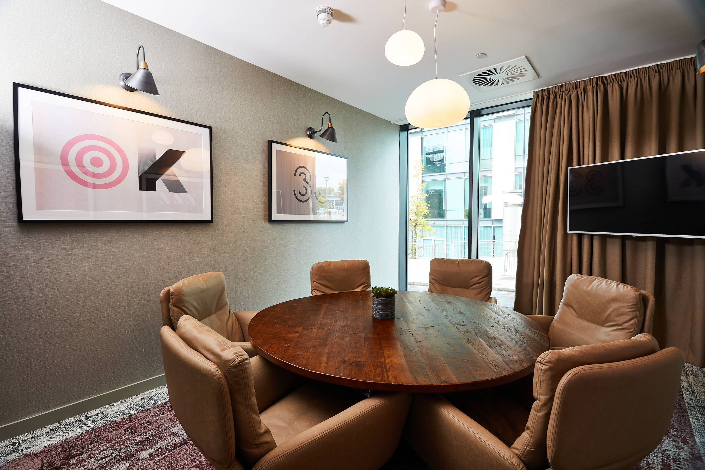 Cozy meeting room on Portland Crescent featuring a round wooden table surrounded by five beige chairs. Walls adorned with abstract art and modern lighting. A large window with curtains provides natural light, while a flat-screen TV is mounted on the wall for presentations.