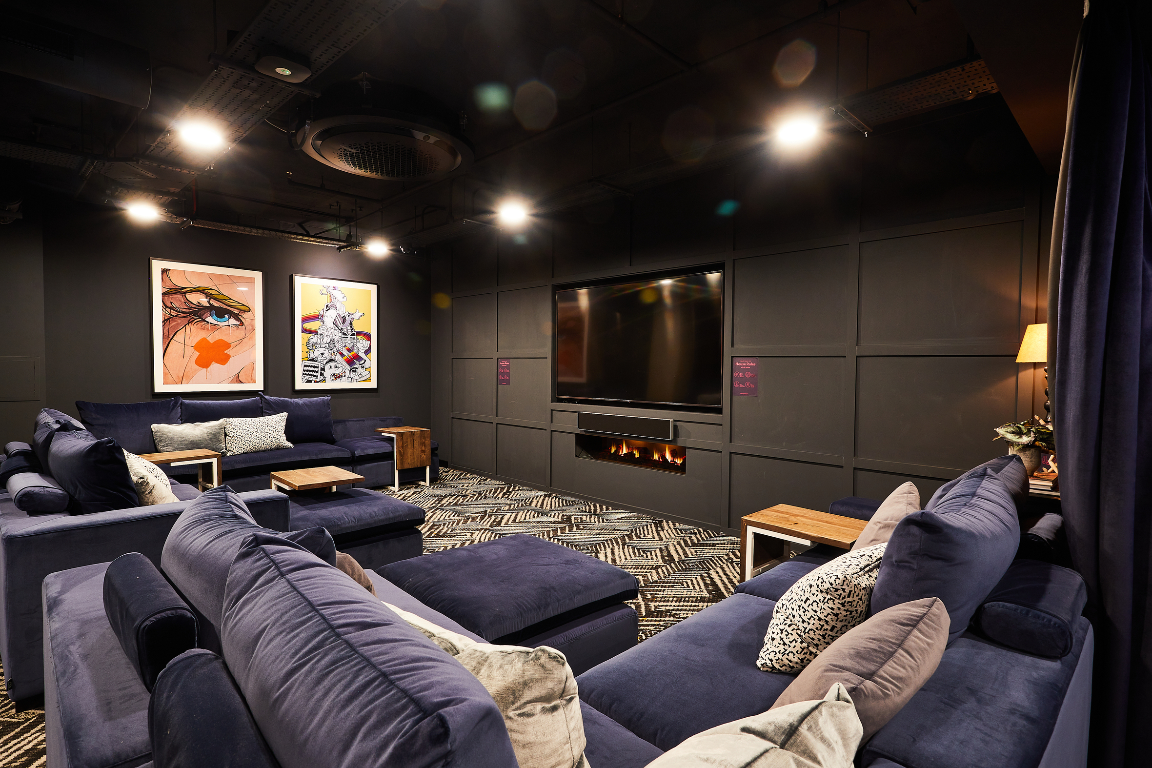 Cozy Portland Crescent home theater room with dark walls and ceiling, featuring navy blue sofas and cushions. Two artworks hang on the wall. A large flat-screen TV and electric fireplace are centered. Soft ambient lighting and patterned carpet add warmth.